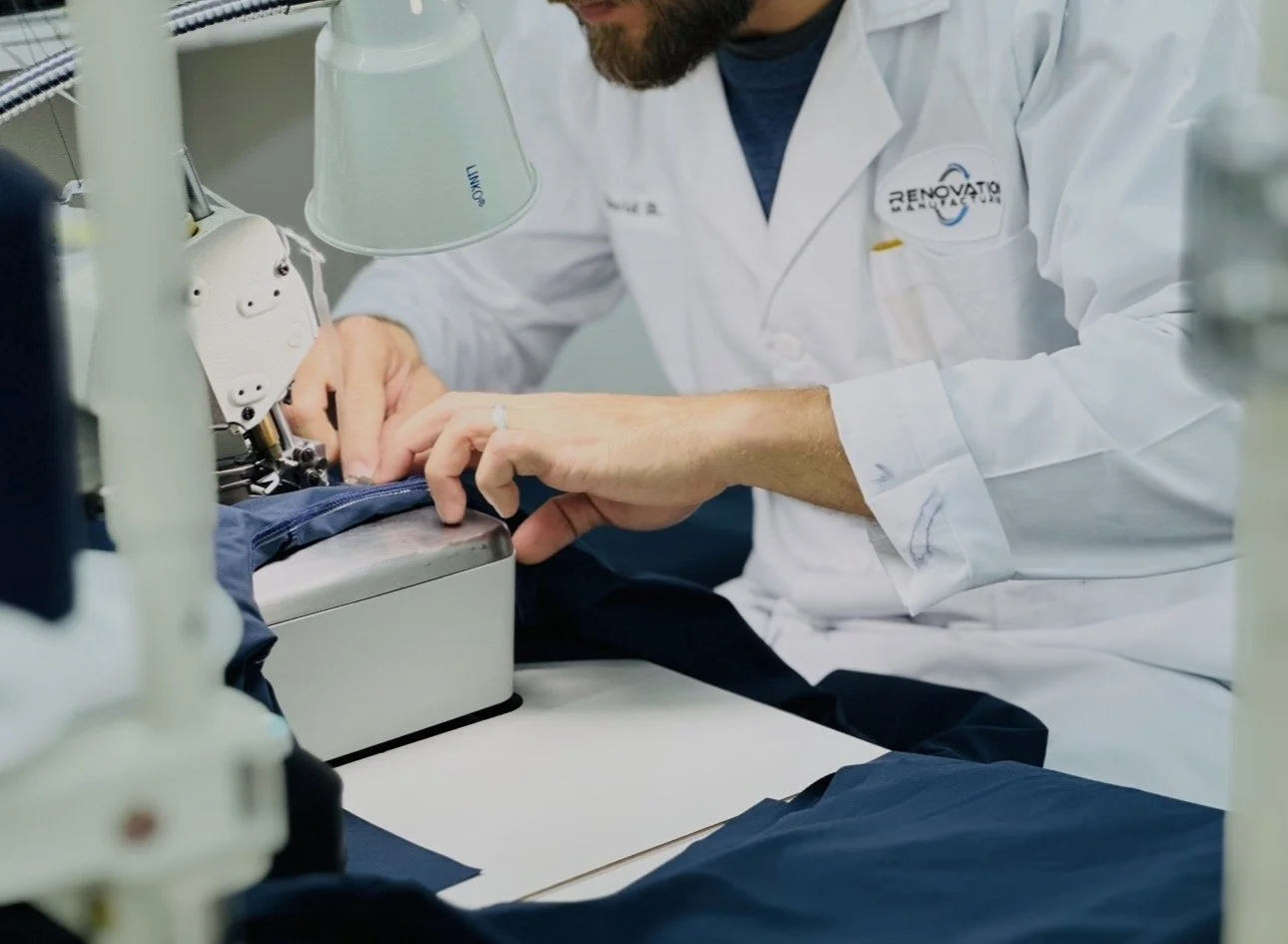 A person working with fabric or sewing machine in a laboratory or workshop setting, wearing a white lab coat with a logo.
