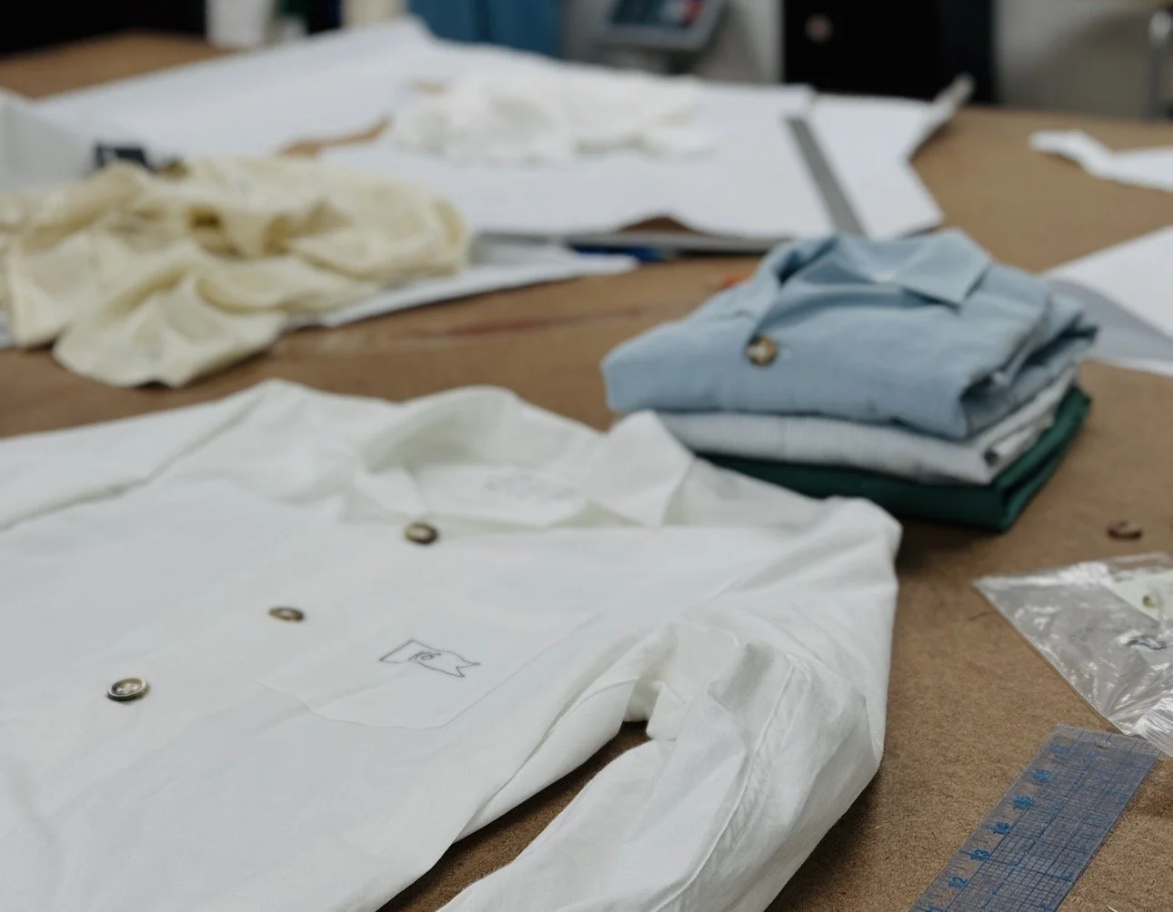Flat lay of sewing workspace with folded shirts, fabric scraps, and sewing tools on a wooden table.