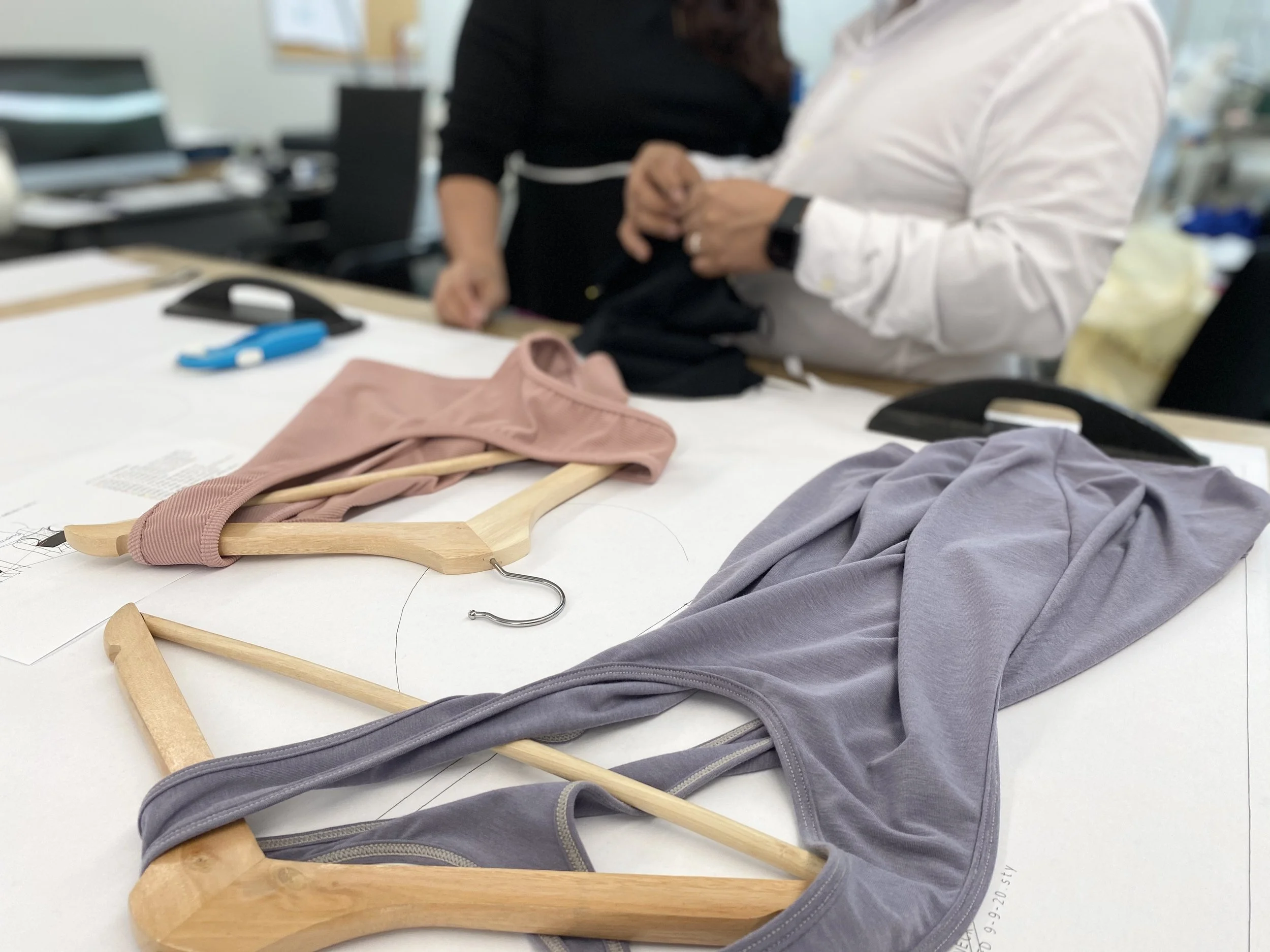 Two women working together at a table with clothing and hangers, one wearing a white shirt and the other wearing a black top.