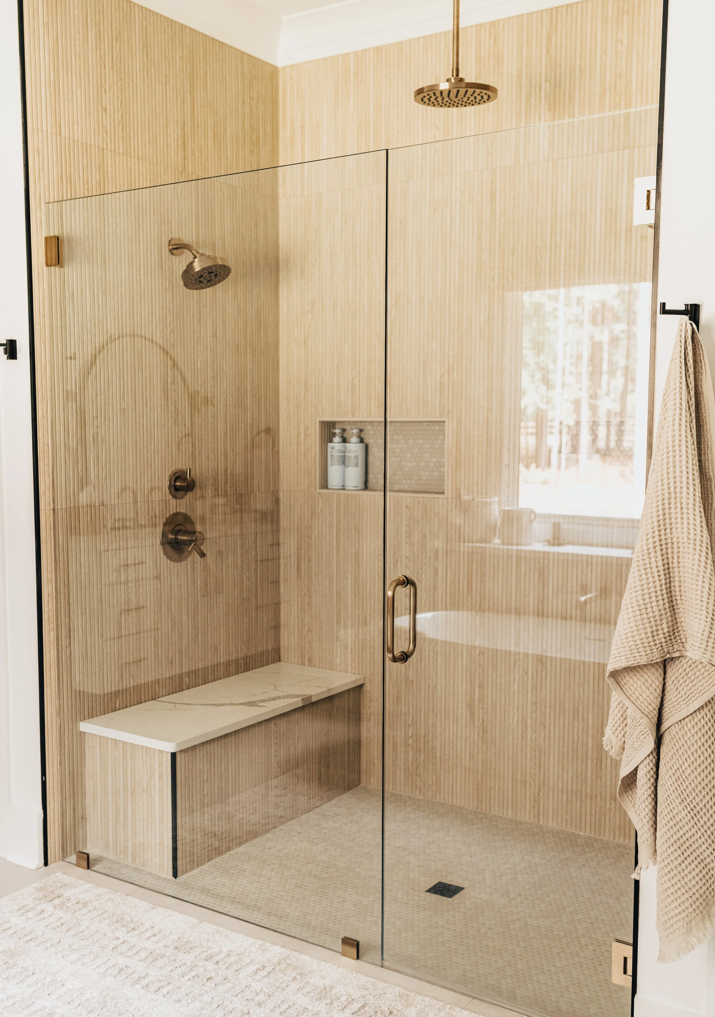 Wood tiled large shower with gold fixtures and a sitting bench with white marble