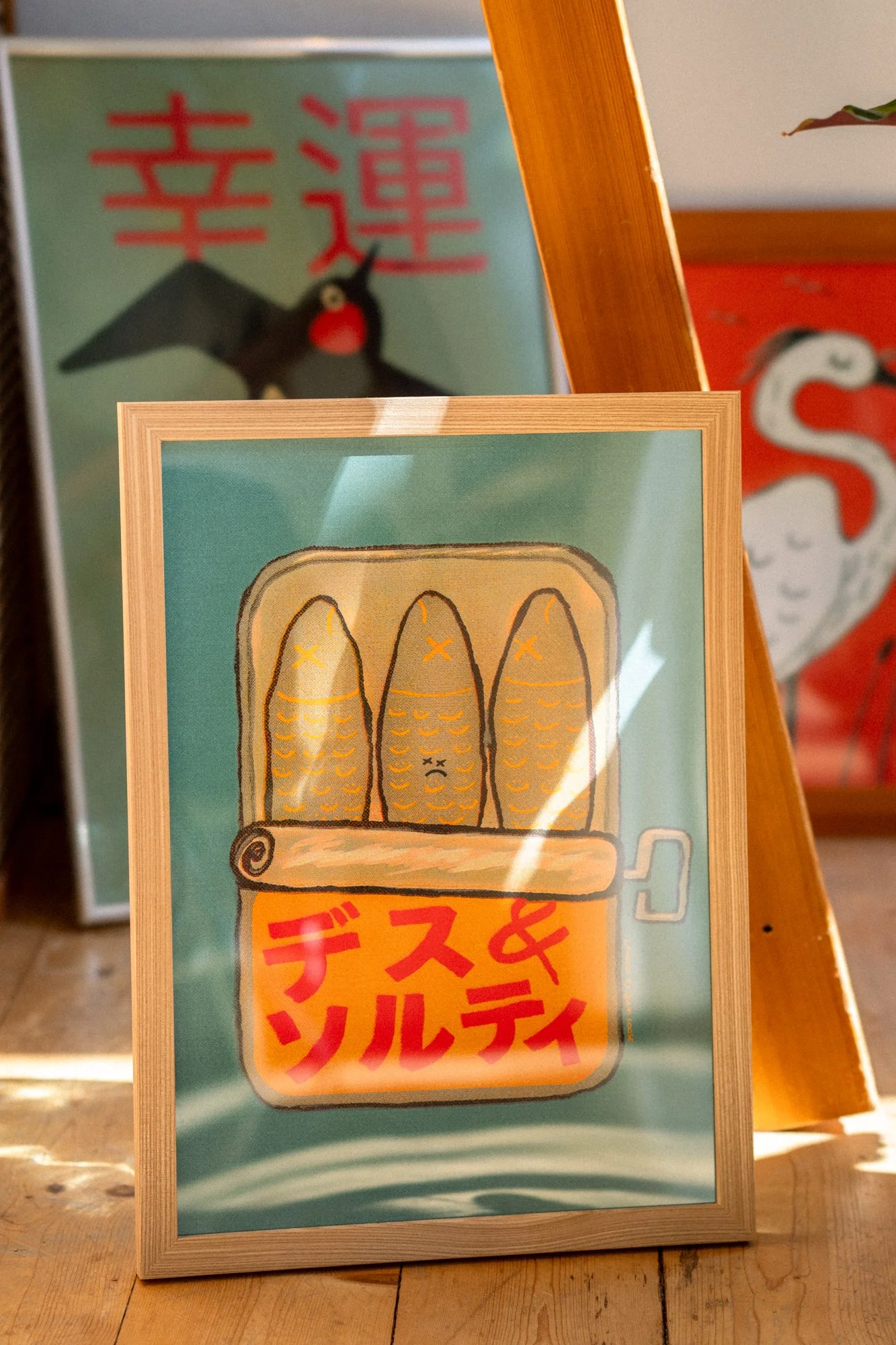 A framed poster with a cartoon illustration of three fish on a book or box, with Japanese text below, placed on a wooden surface. In the background, there are two other posters, one featuring a black bird with a red beak and Japanese characters, and 