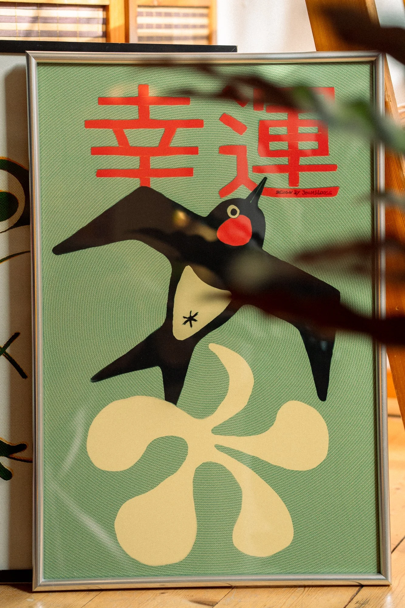 A framed artwork with a green background, featuring a cartoon-style black and white bird with a red cheek and a long beak, along with red Chinese characters and a yellow floral design.