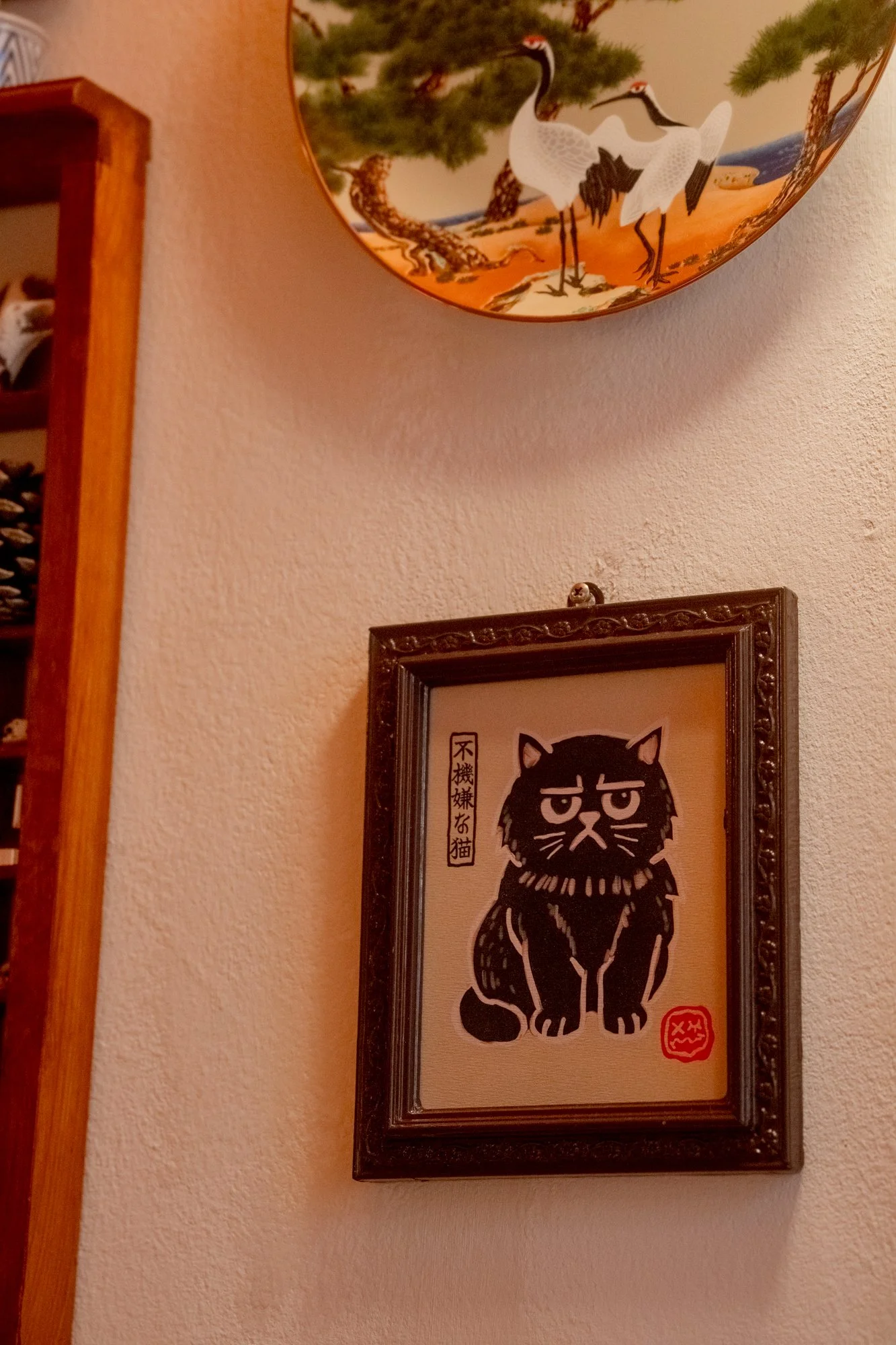 A framed black drawing of an annoyed-looking cat with Japanese characters and a red stamp on the wall, with a decorative plate of cranes and a pine tree in the background.