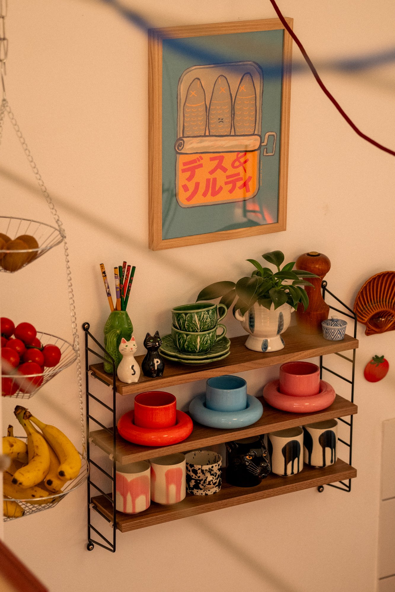 Decorative kitchen shelf with assorted ceramic bowls, cups, and figurines, including a white cat, black cat, and black panther, with a potted plant on top. A framed art piece with a cartoon illustration and Japanese text hangs above the shelf.