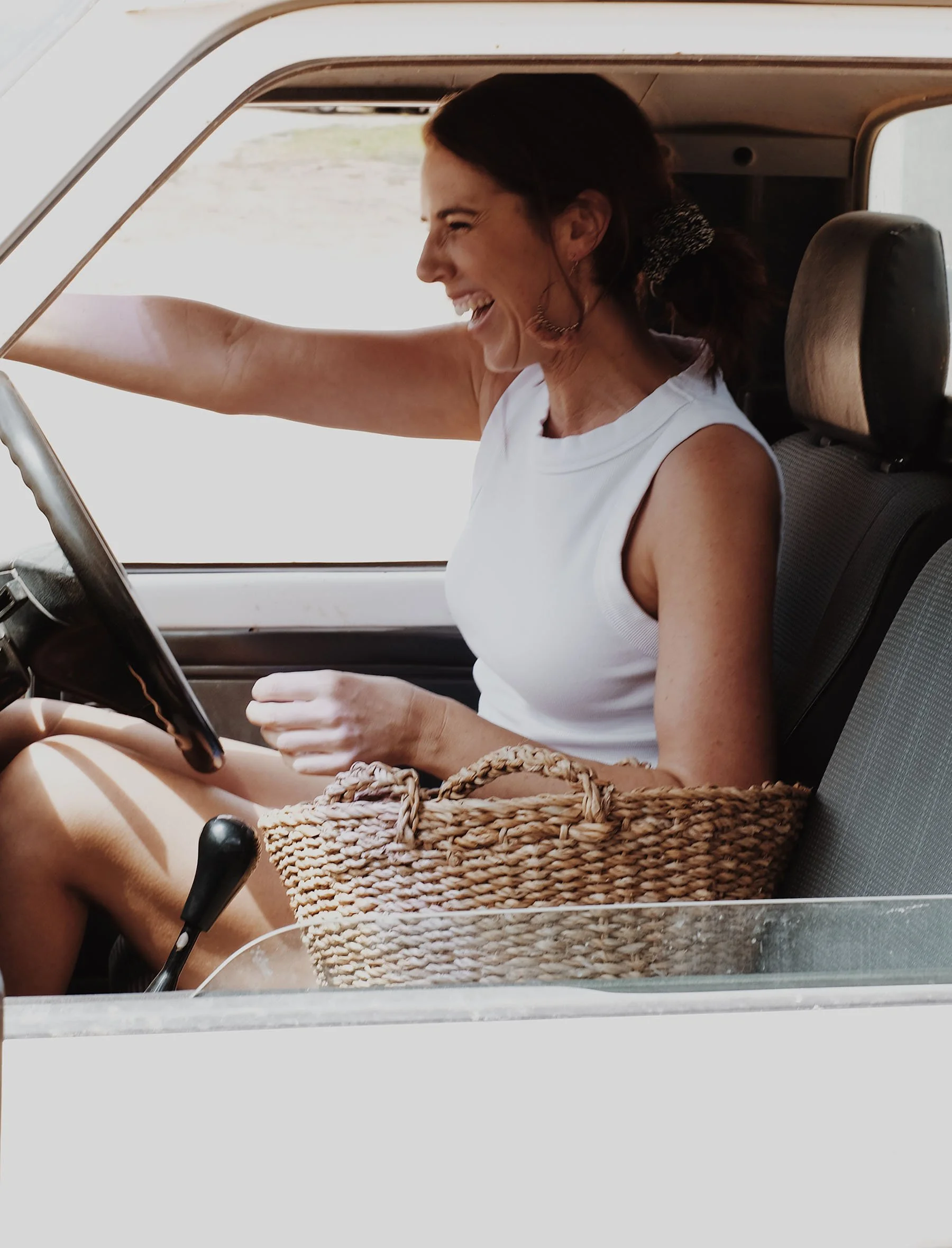 A woman in a white sleeveless shirt sitting in a vehicle, smiling and laughing while holding the steering wheel, with a woven bag on her lap.