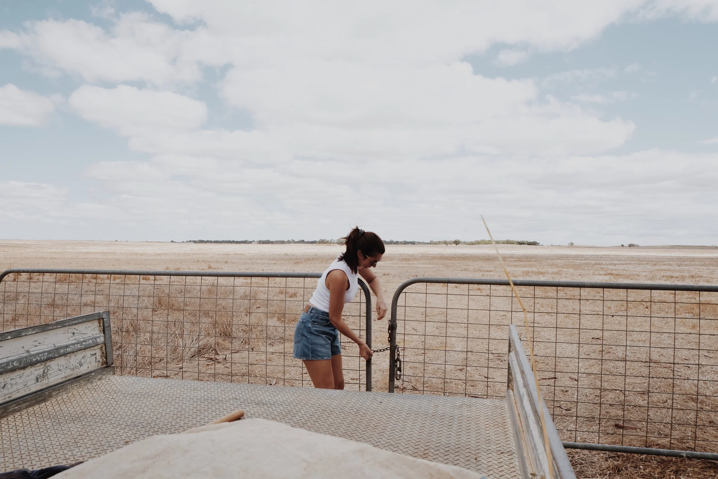 Woman in white tank top and denim shorts unlocking a metal gate on a fence in a dry, open field under a cloudy sky.