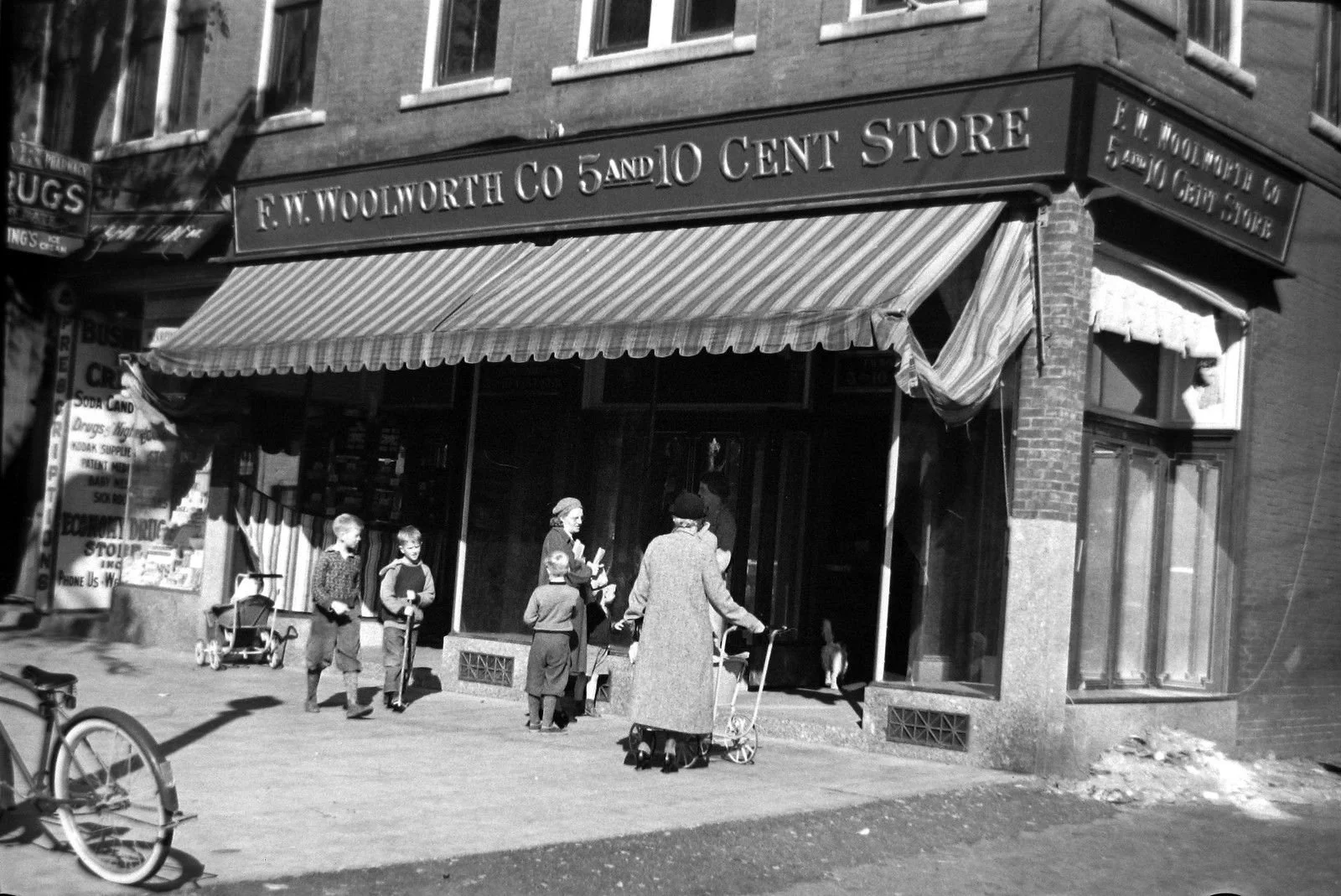 Step back to downtown Natick when a nickel went far! The F.W. Woolworth Co. 5 &amp; 10 at 16 W. Central St.&mdash;open by 1921 (per the Natick Bulletin)&mdash;stands under its striped awning with kids on scooters, a woman beside a pram, and a bicycle