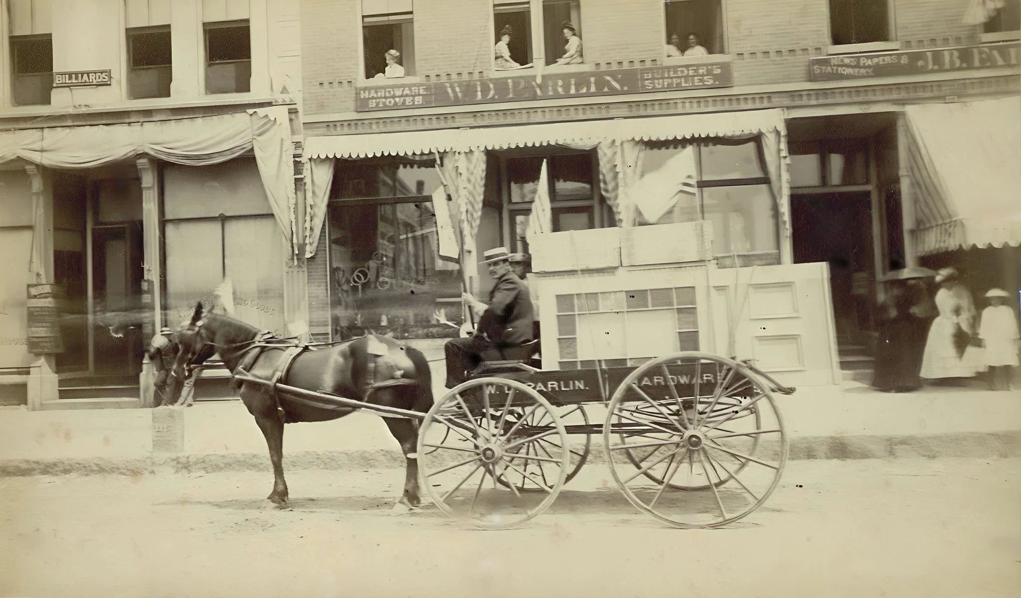 20 Main St., a horse-drawn wagon for W. D. Parlin Hardware waits outside the store, where hardware, stoves, and building supplies were delivered around town by horse and wagon. Look closely&mdash;you can spot people watching from the windows above!

