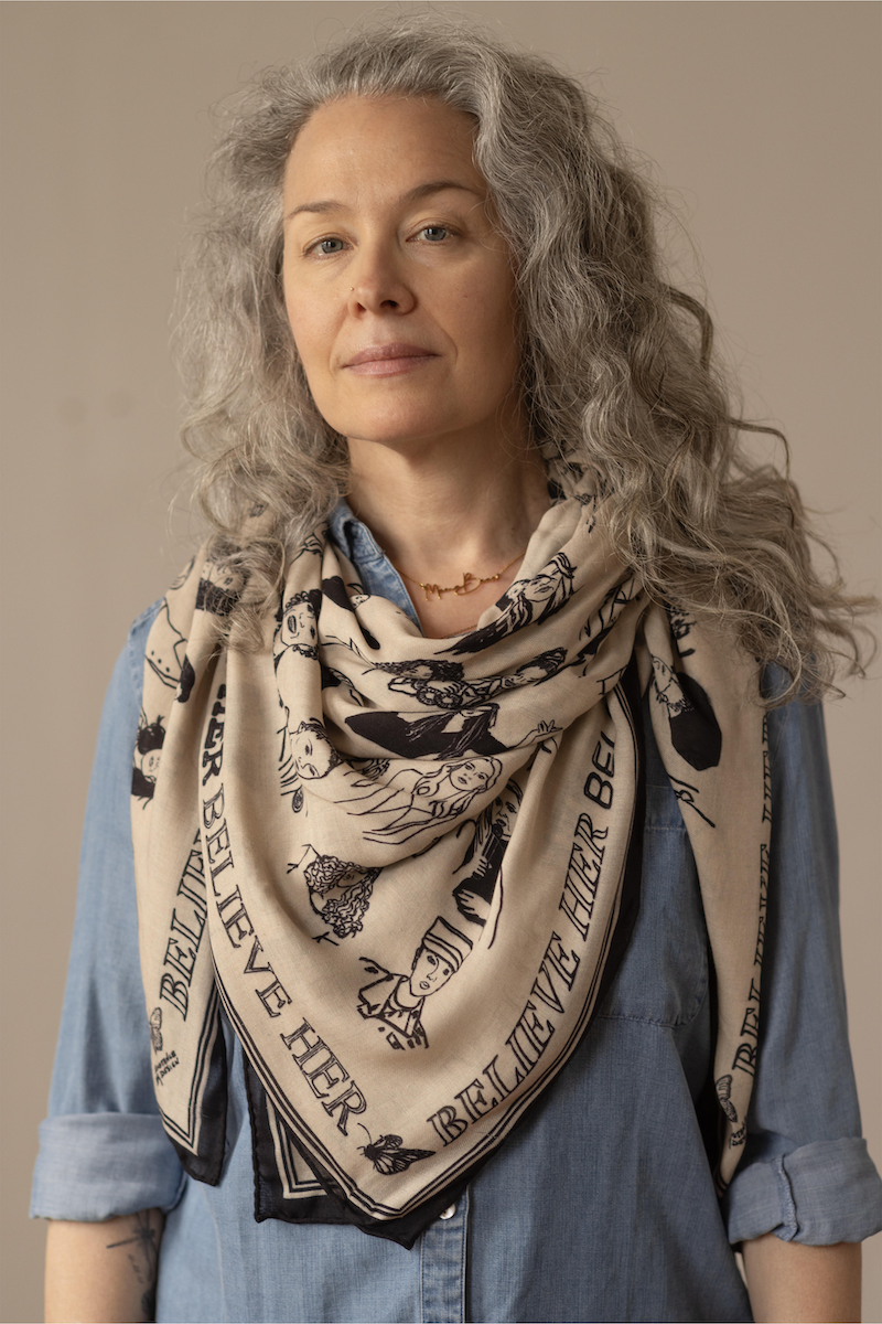 A woman with gray, curly hair wearing a beige scarf with black illustrations and text, layered over a denim shirt, against a neutral background.