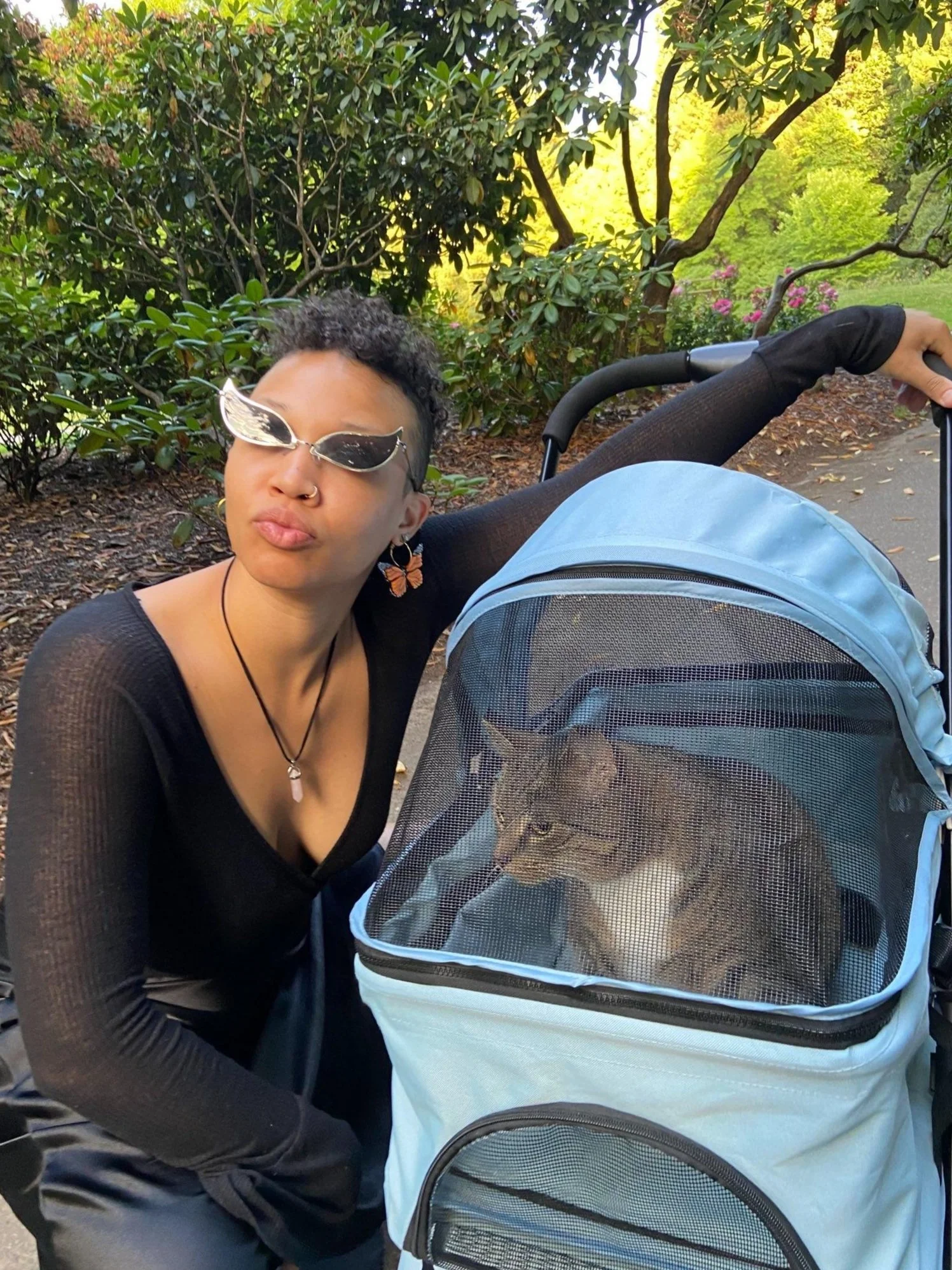 A picture of Lovey in a park pursing its lips playfully the sun glinting off its mirror sunglasses, butterfly hoop earrings, and crystal hanging around its neck. Squatting and draping their arm over the handle of a stroller with their cat Luna inside