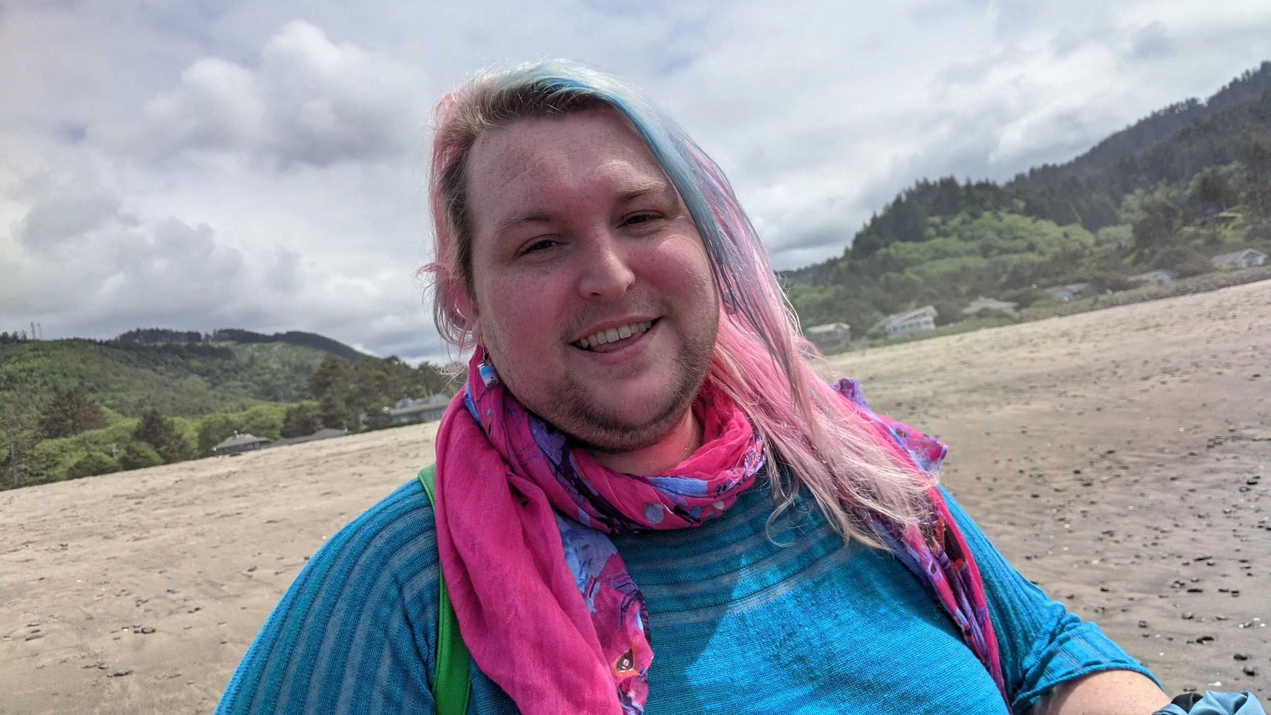 A picture of Princess smiling on the beach on a cloudy day. She has rainbow pastel hair, a colorful silk neckscarf, and curious quandaries on the mind