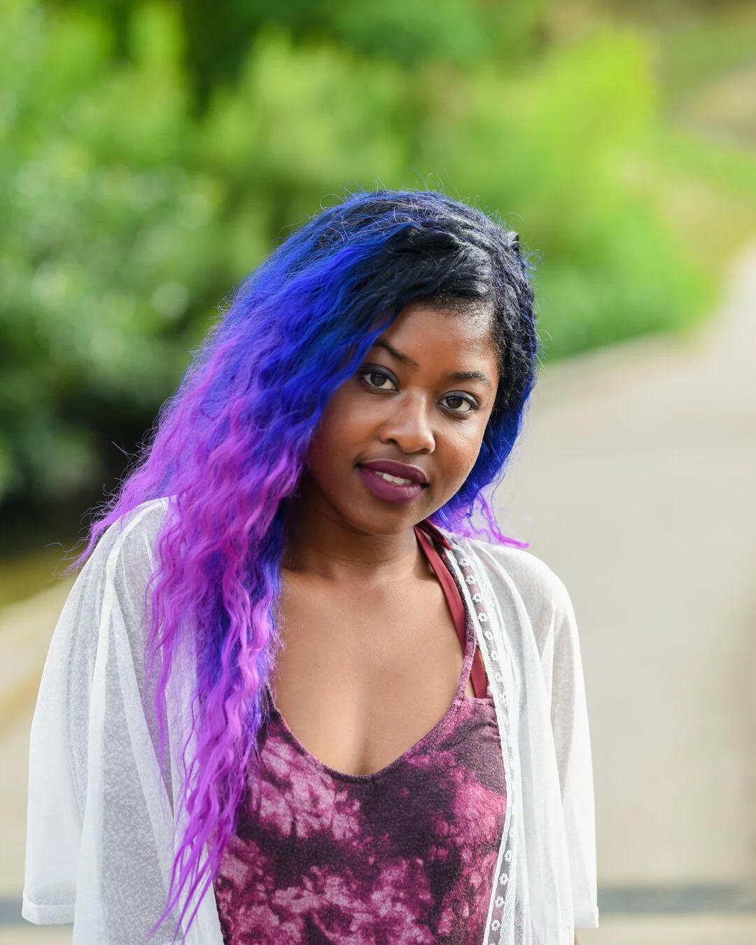 A woman with long, wavy hair that fades from black to vibrant purple and blue, standing outdoors with green foliage in the background.