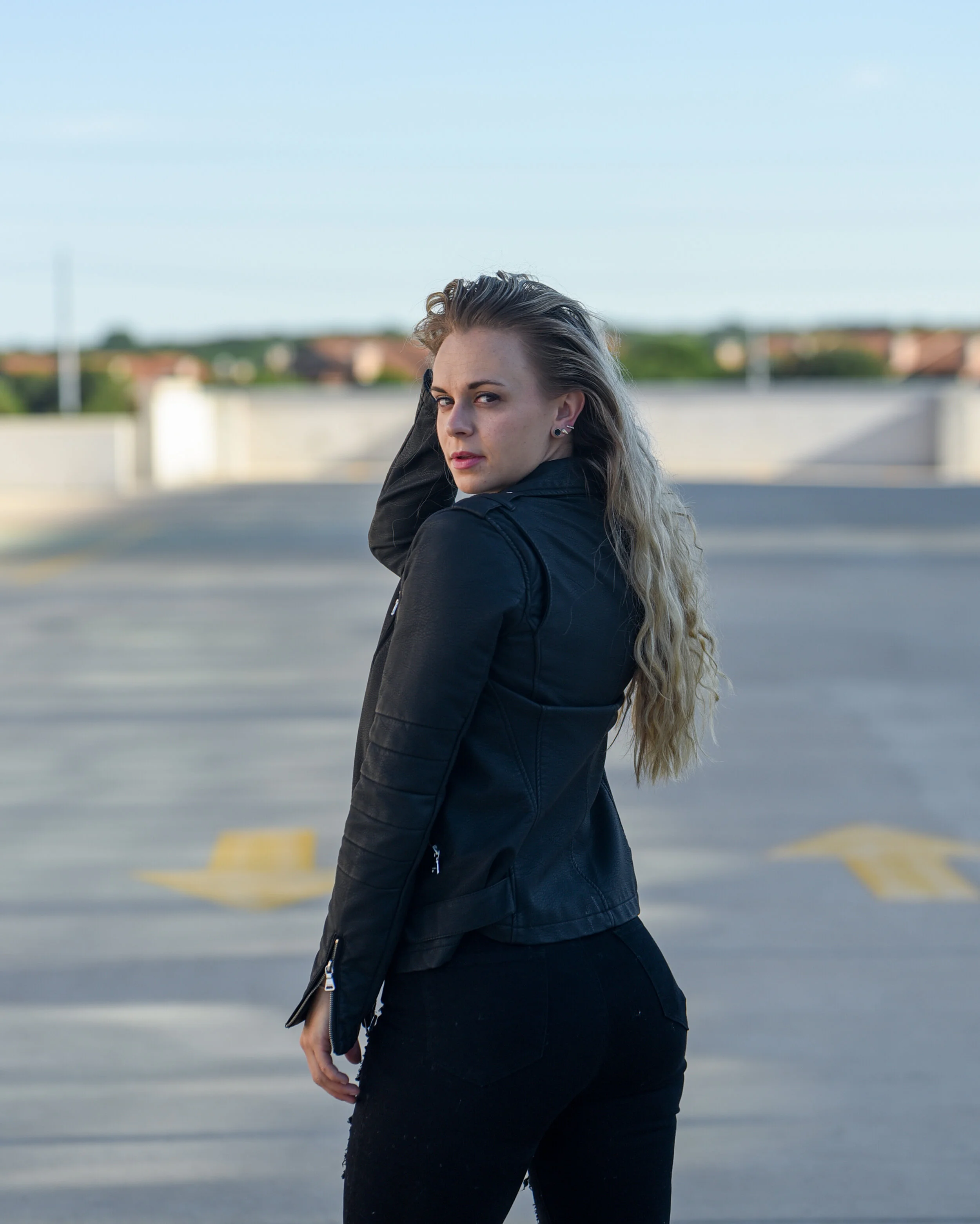 A woman with long wavy blonde hair and fair skin, wearing a black jacket and black jeans, standing outdoors against a blurred background of a parking lot and suburban houses, looking back towards the camera with a serious expression.