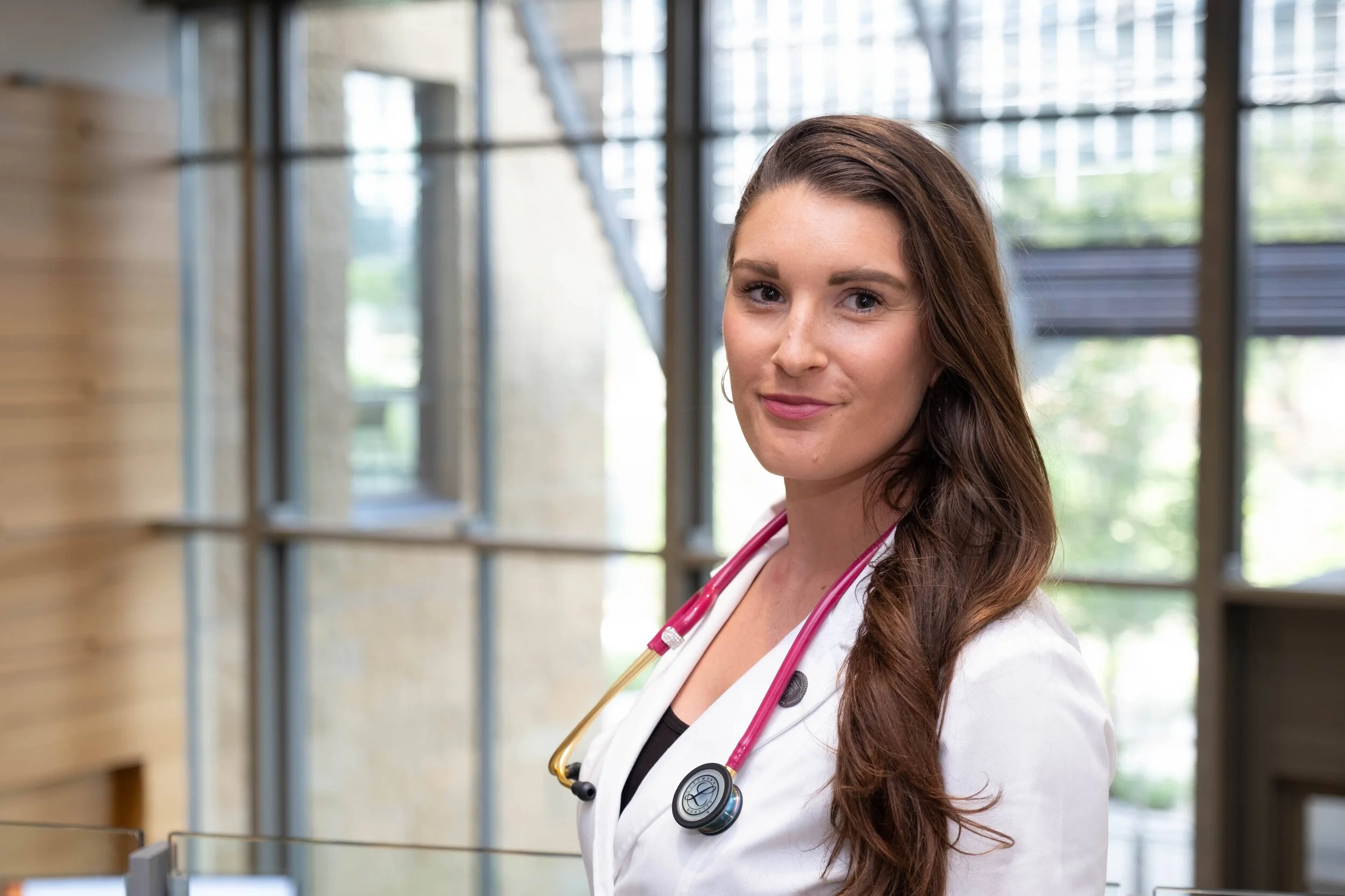 A young female healthcare professional with long brown hair, wearing a white coat and a pink stethoscope around her neck, smiling in a modern medical facility with large windows.