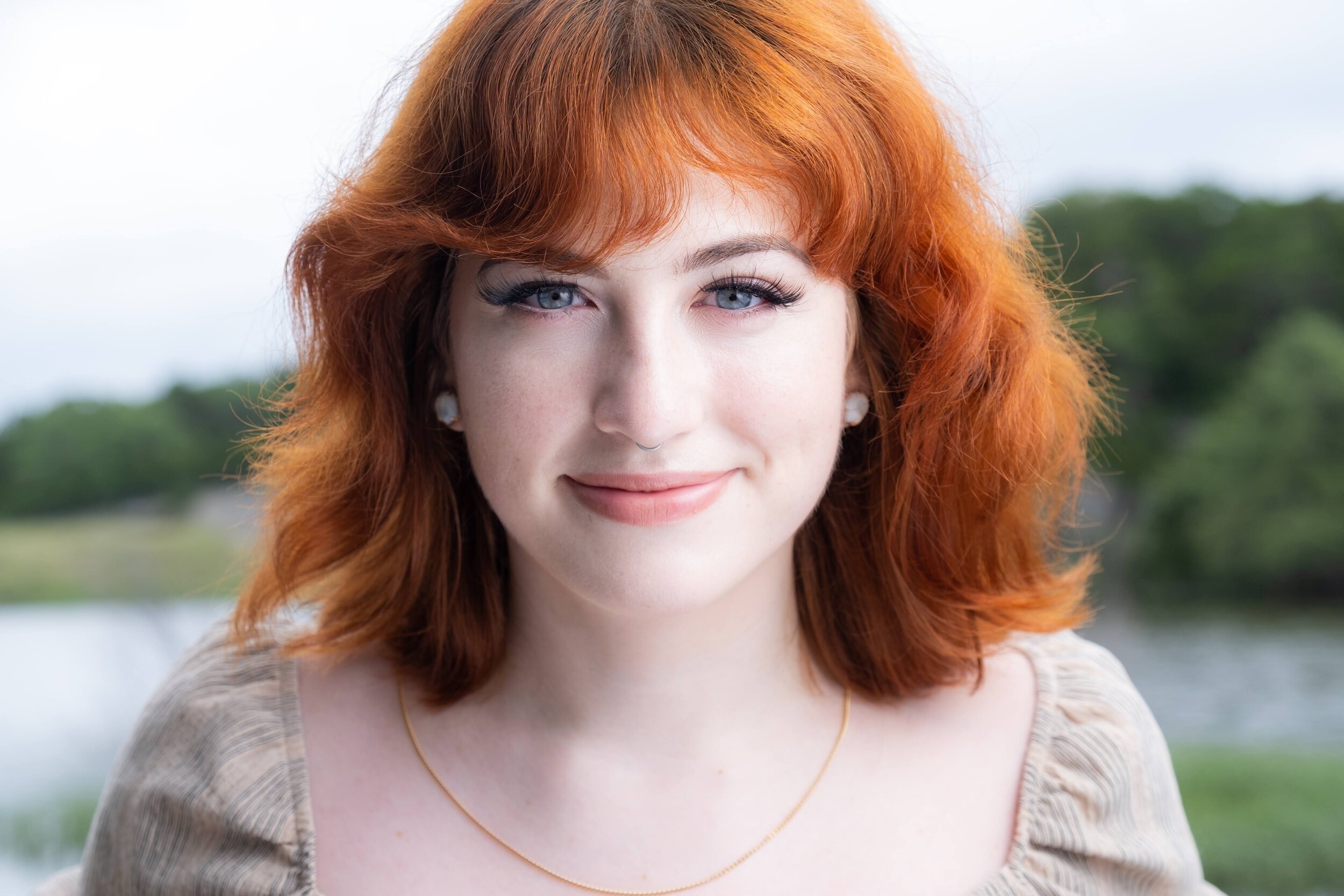 A young woman with red hair and blue eyes smiling outdoors with a blurred green foliage background.