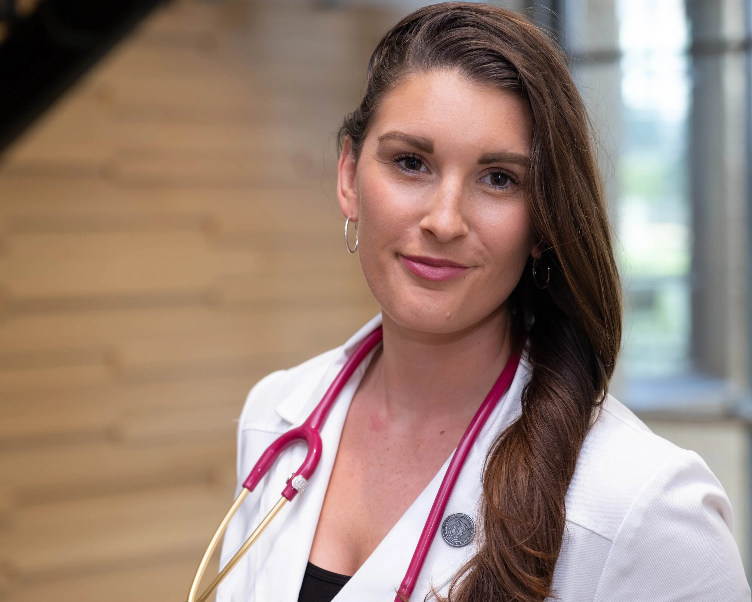 A female healthcare professional, possibly a nurse or doctor, with long wavy brown hair wearing a white lab coat and a pink stethoscope around her neck, smiling softly in a professional setting with a blurred background.