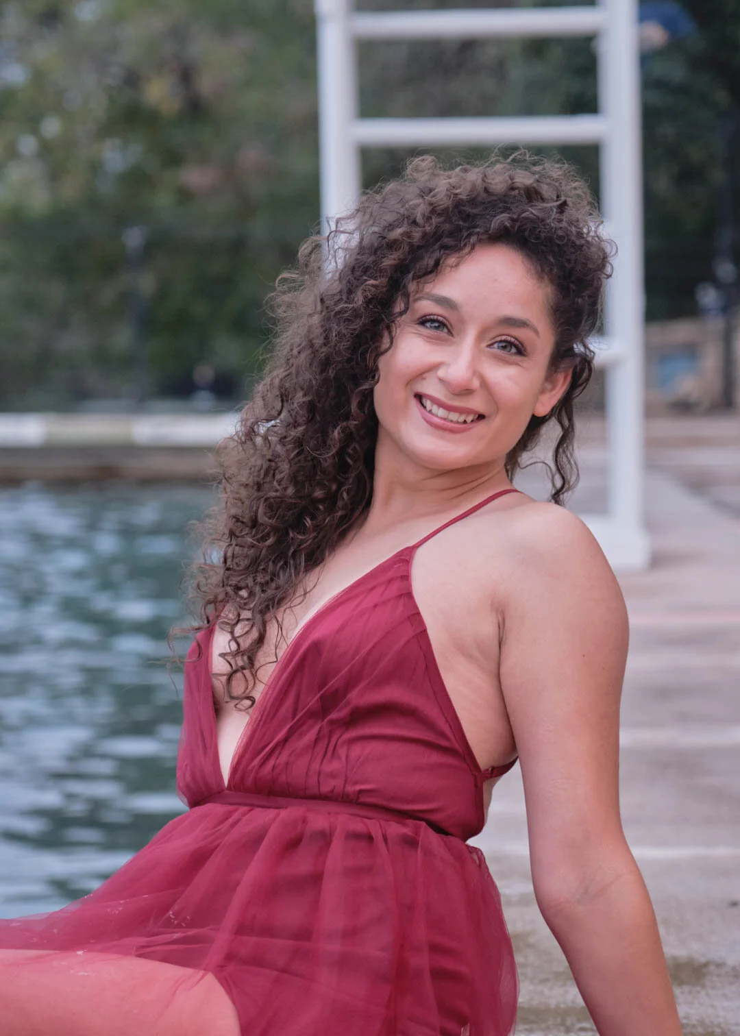 A woman with curly hair and a red dress sitting by a body of water near a dock or pier, smiling at the camera.