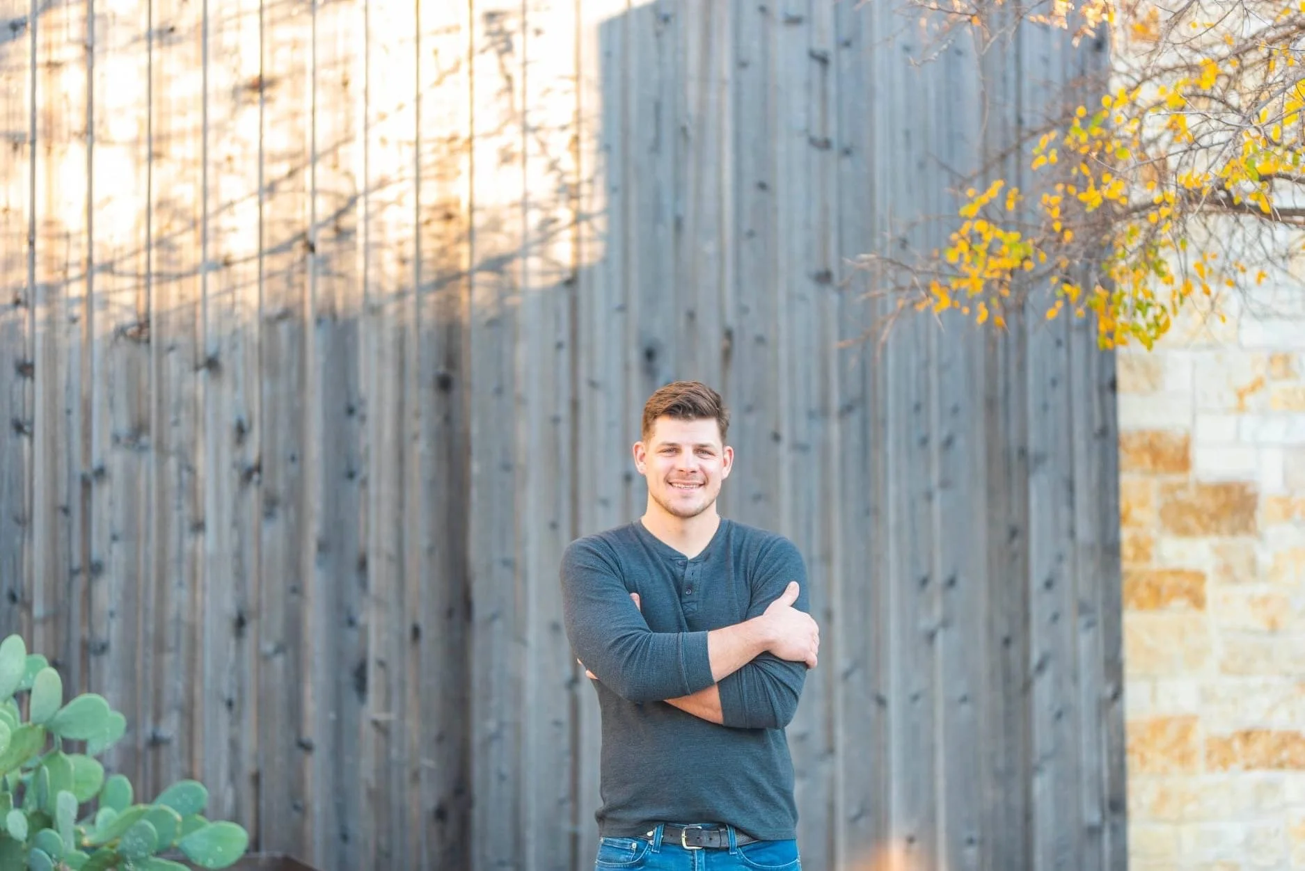 A smiling man with crossed arms standing outdoors in front of a wooden fence, wearing a long-sleeve dark gray shirt and jeans.