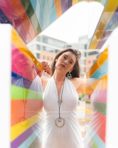 Woman in a white dress looking into a colorful rainbow tunnel structure outdoors.