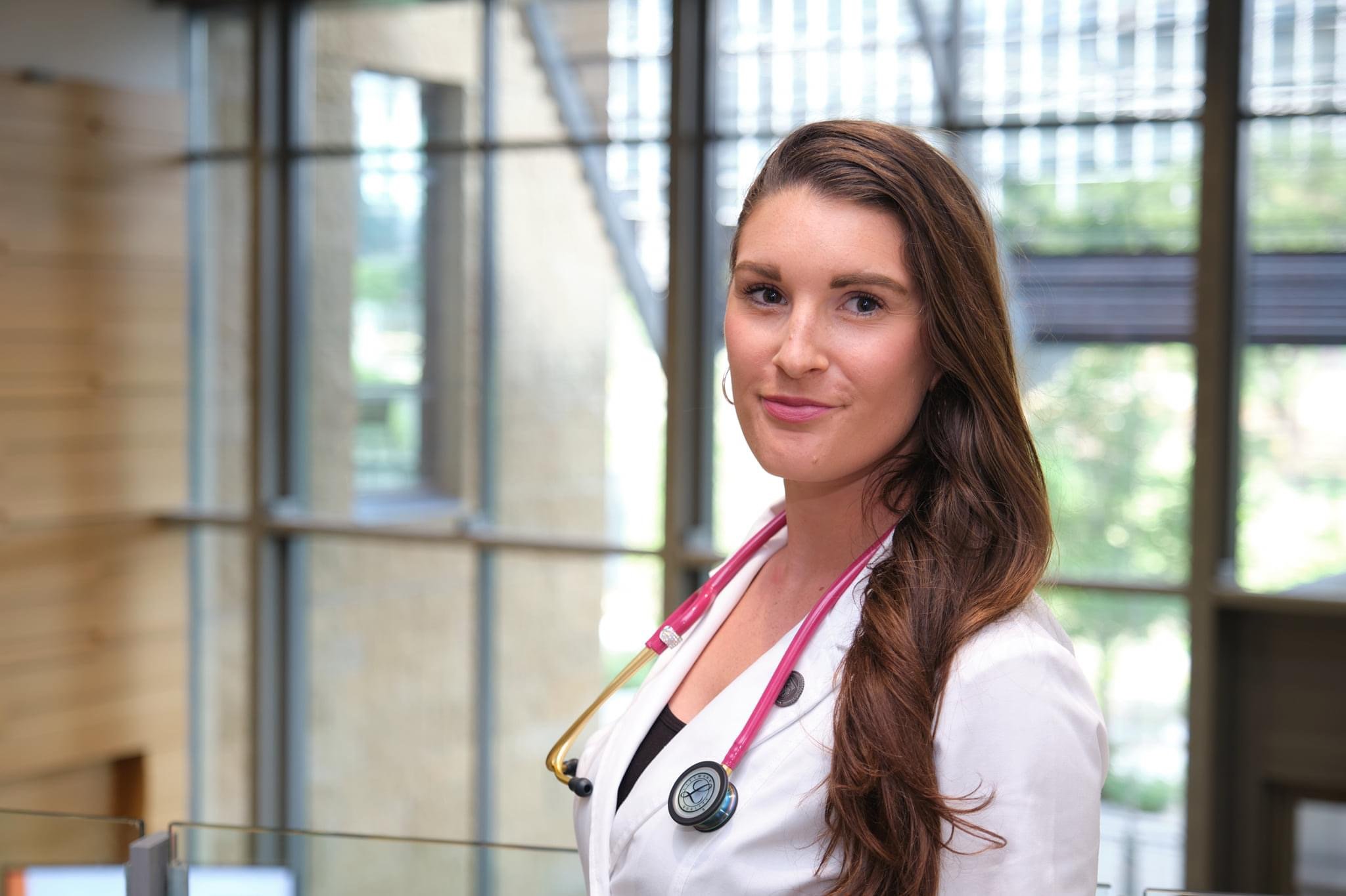 A young female healthcare professional wearing a white coat with a pink stethoscope around her neck, standing in a modern medical facility with large windows and wooden accents.