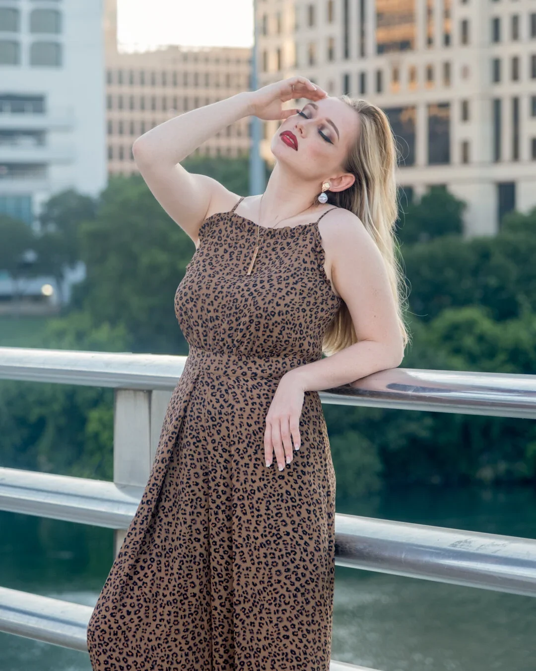 A woman with long blonde hair wearing a leopard print dress and earrings, posing with her eyes closed and one hand on her forehead, leaning on a railing at an outdoor location with tall buildings and greenery in the background.
