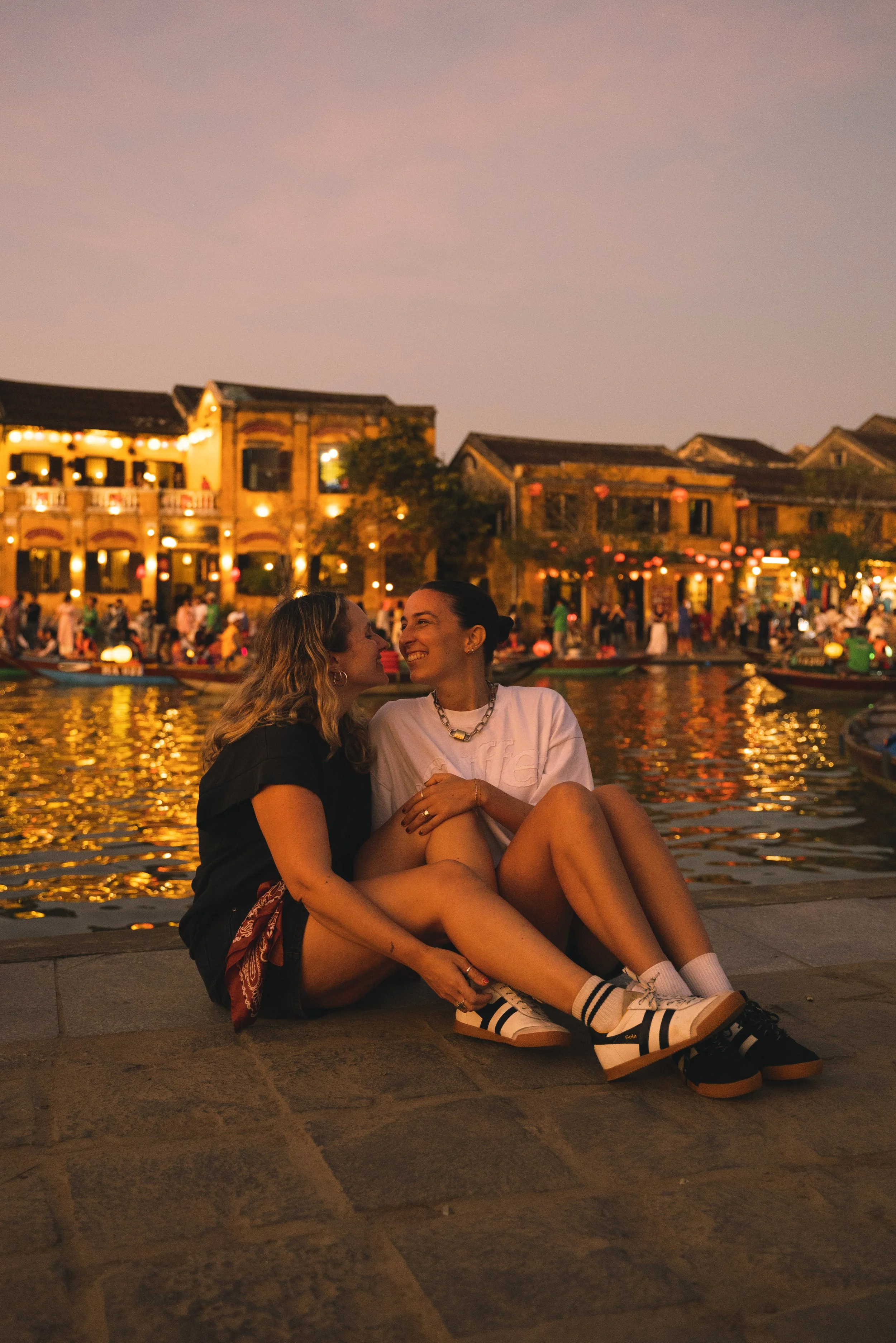 two girls sitting on river while lantern boats go behind them in hoi an vietnam