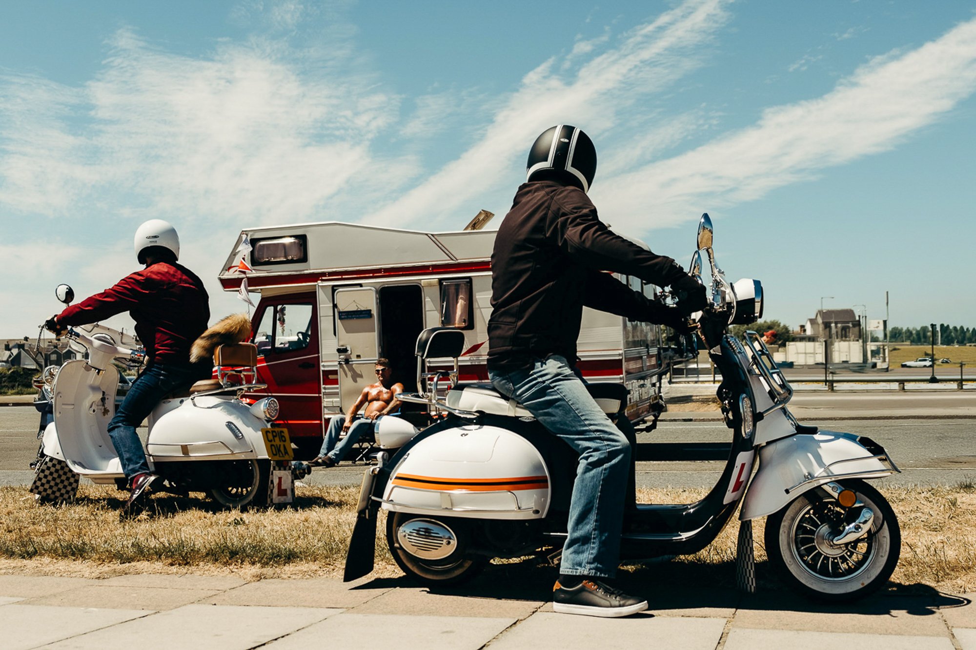 A shirtless man sits outside his camper van, between two people on vespas on a sunny day at coney beach, bridgend. 