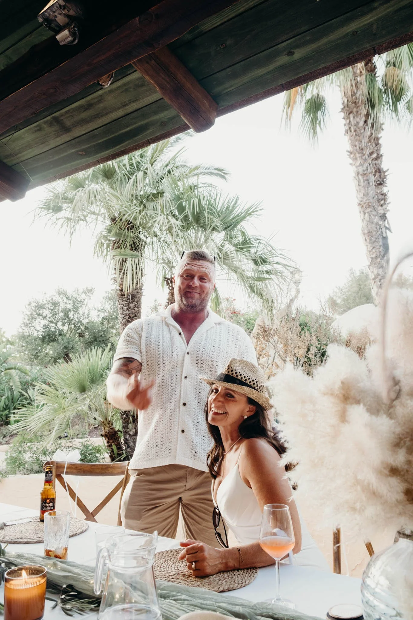 A man and woman at an outdoor table, smiling and enjoying a tropical setting with palm trees, some beverages, and decorative plants.