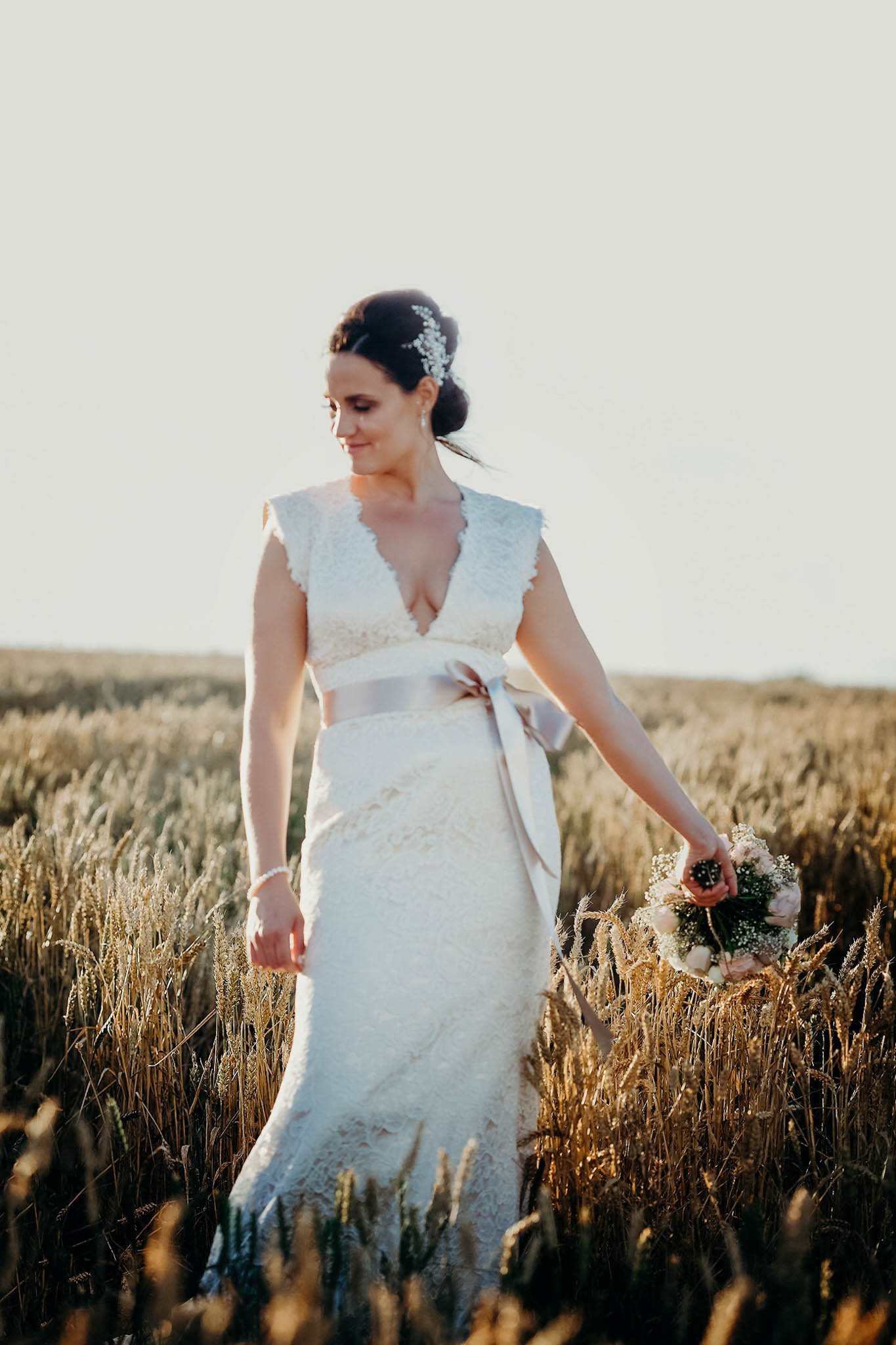 A bride in a white lace wedding gown holding a bouquet walking through a wheat field during sunset.