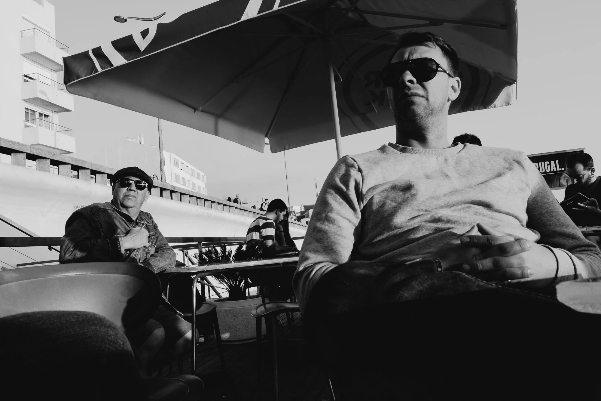 Black-and-white photo of three men seated at an outdoor café table under large umbrellas on a seaside promenade. The man in the foreground wears sunglasses and rests casually, while others drink and converse. Modern buildings and a "PORTUGAL" sign ar