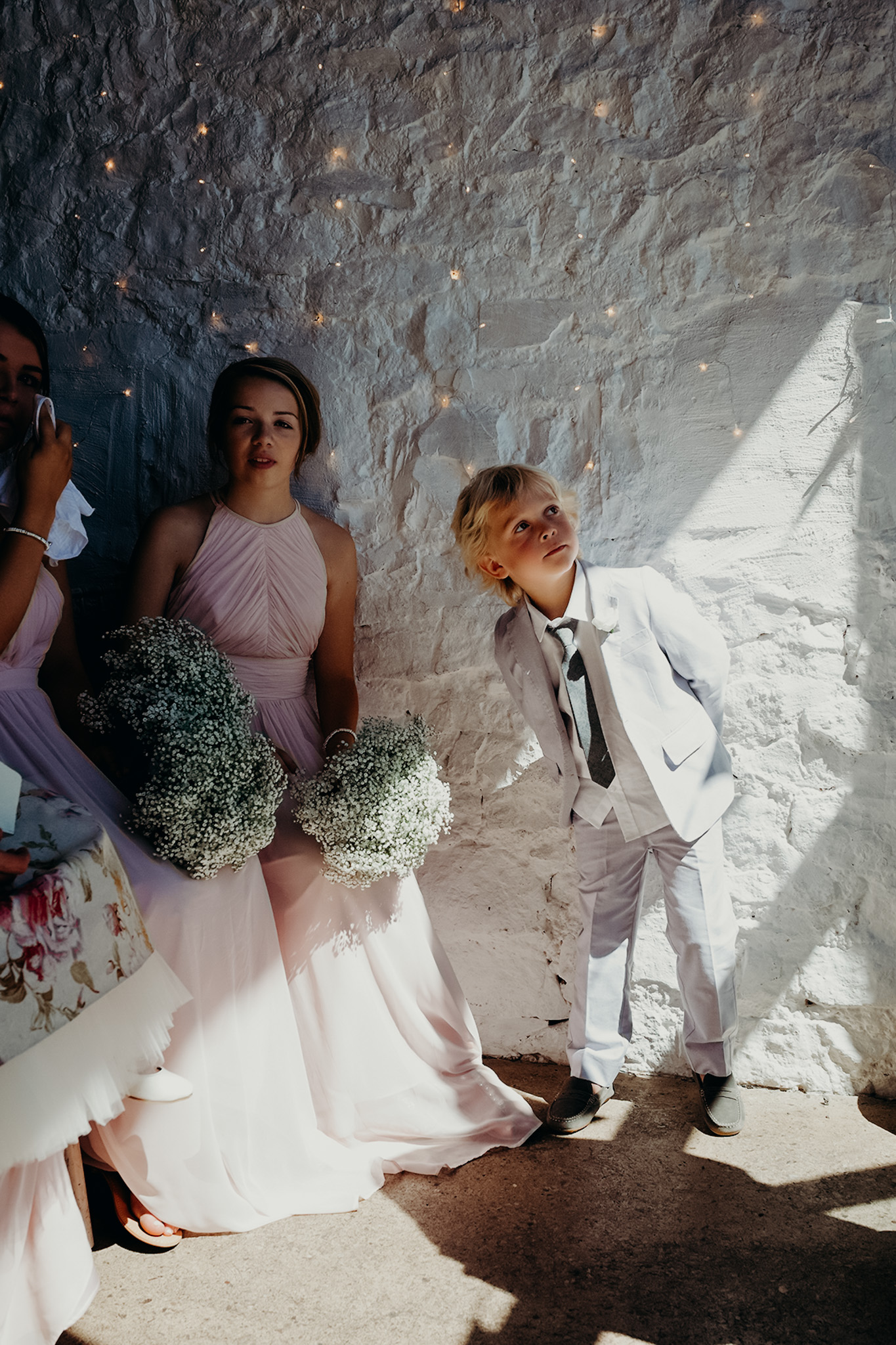 A group of people, including a young boy and two women in pink dresses holding bouquets, standing against a white stone wall decorated with small lights, during a wedding in bridgend. Shot by Jason Evans photography Bridgend
