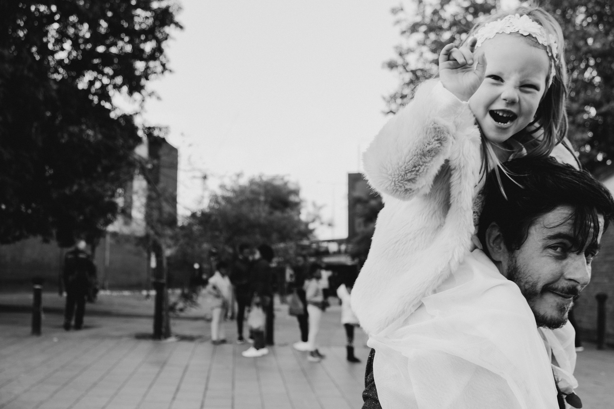 A man carries a young girl on his shoulders in an outdoor wedding in London. The girl is smiling and making a peace sign with her hand. Shot by Jason Evans photography Bridgend