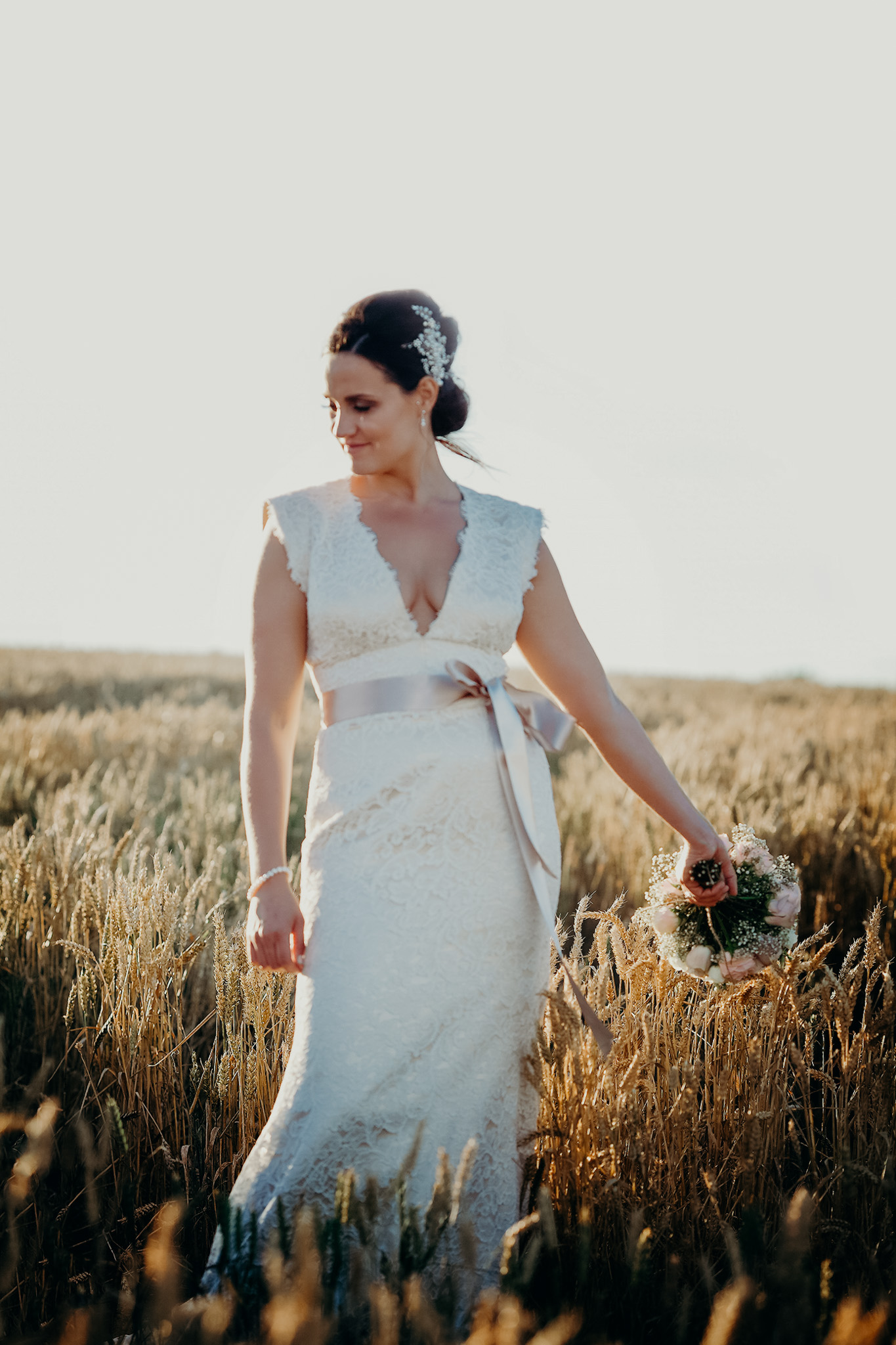 Bride in a white lace wedding dress holding a bouquet of flowers, standing in a wheat field during sunset at a wedding in llantwit major. Shot by Jason Evans photography Bridgend