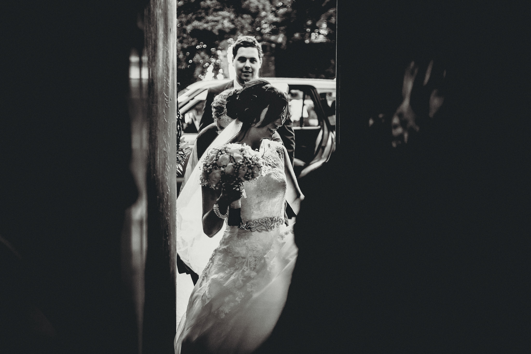 A bride in a wedding dress holding a bouquet, smiling and looking down, as she leaves a vehicle, with a groom and guests visible in the background at fon mon castle. Shot by Jason Evans photography Bridgend