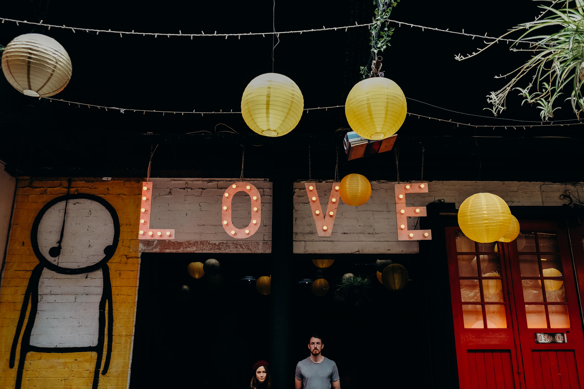 Decorative hanging paper lanterns and a sign spelling 'LOVE' with lit bulbs hanging above two people standing in front of a building with a red door, a mural of a cartoon figure, and greenery. Shot by Jason Evans photography Bridgend
