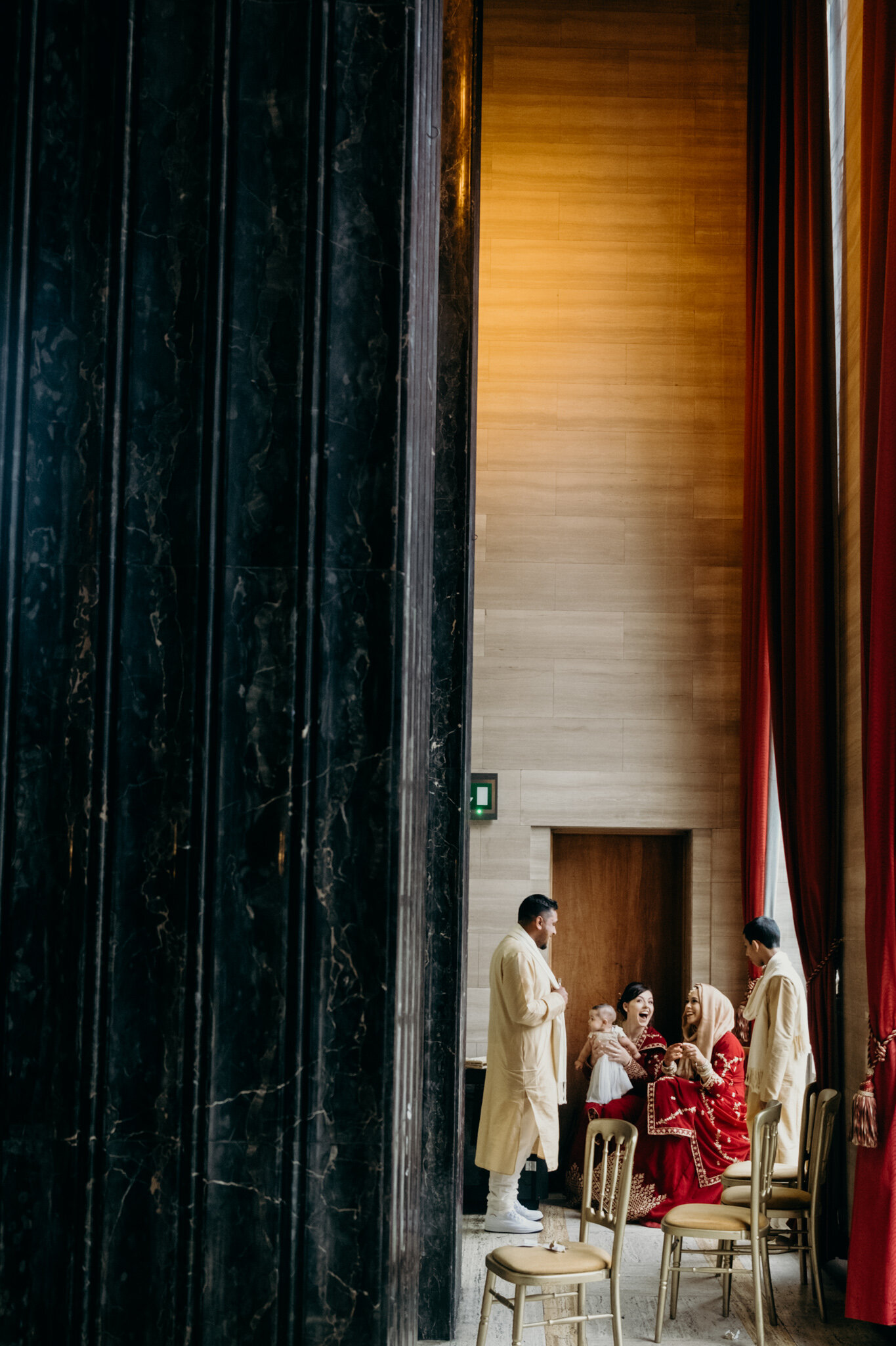 People gathered in a formal room with high ceilings, red curtains, and wooden walls, engaged in conversation, with one person holding a baby at a wedding in Cardiff. Shot by Jason Evans photography Bridgend