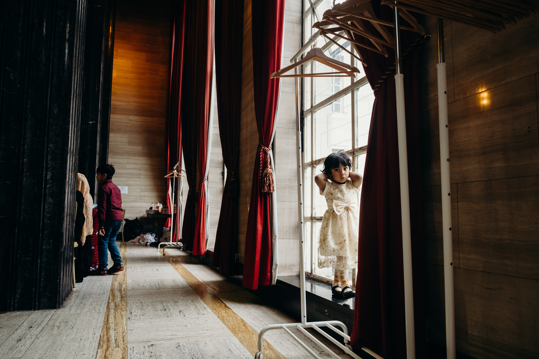 A young girl in a white bridesmaid dress with a bow stands behind a fitting room curtain, holding her hair back and looking at the camera, with red velvet curtains and a wooden wall in the background. Shot by Jason Evans photography Bridgend