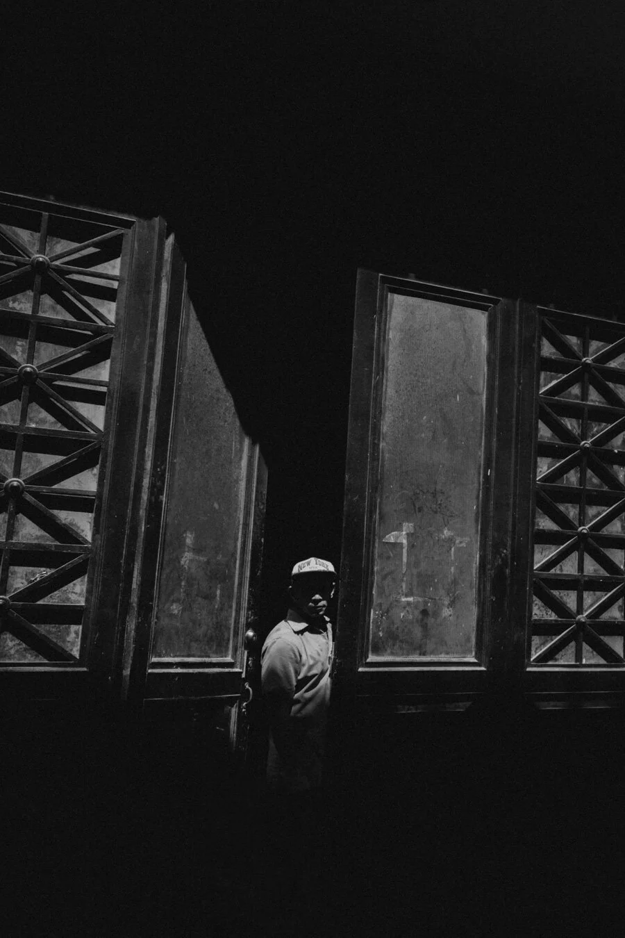 A person standing behind large open double doors, partially visible, with high contrast black and white lighting in Havana, Cuba. 