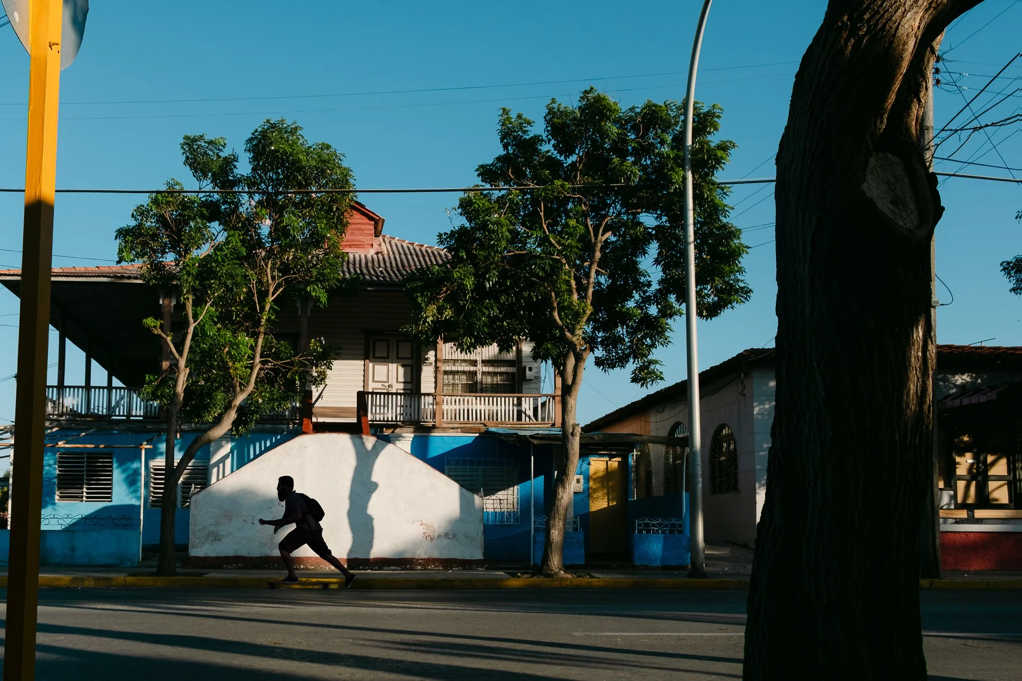 A man skateboards down an empty residential street in late afternoon sunlight, casting a long dramatic shadow on a white wall. Colorful low-rise houses with tiled roofs and trees line the street in Cuba under a clear blue sky.