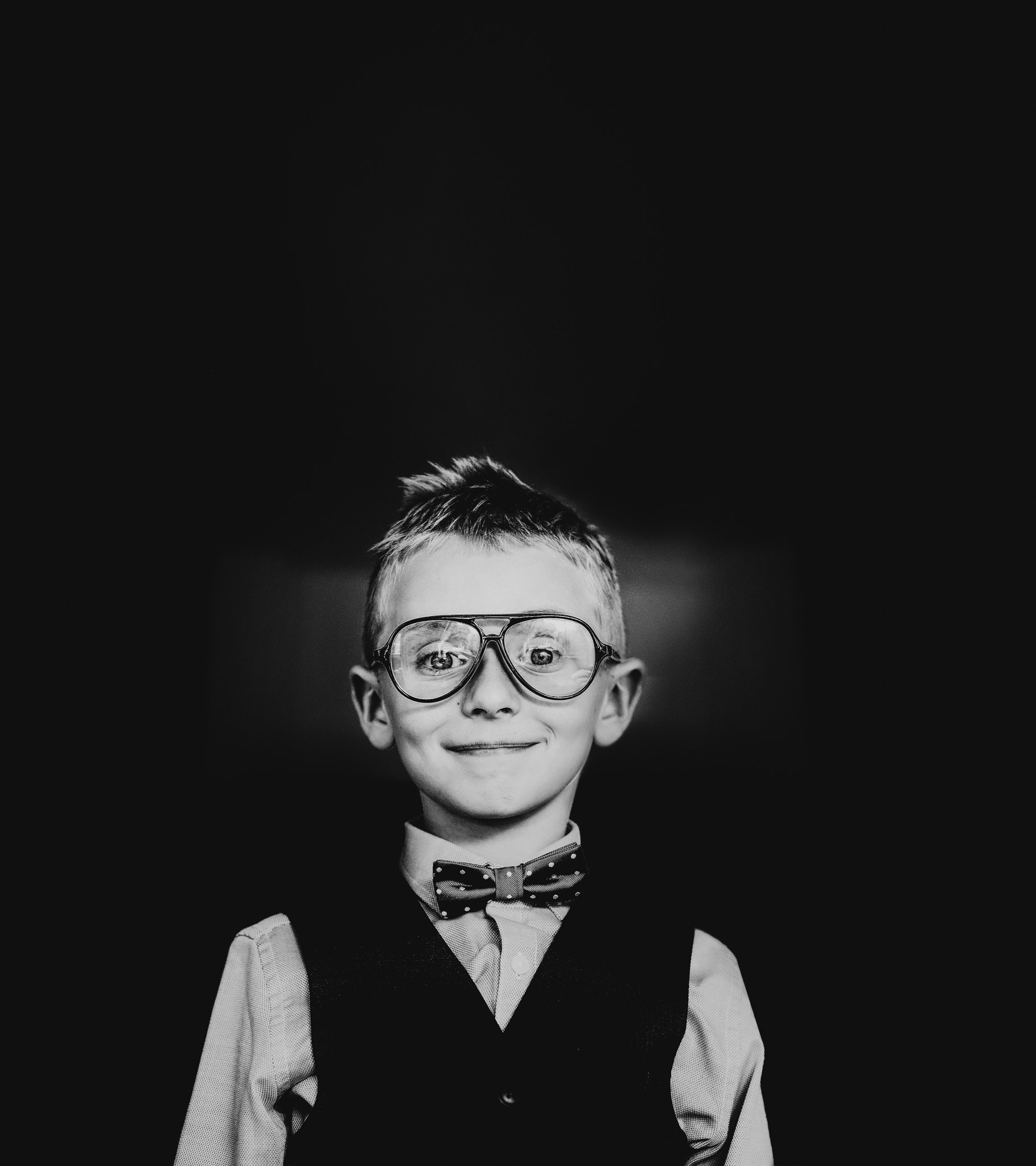 Black and white photo of a young boy with glasses, wearing a bow tie and vest, smiling at the camera. Shot by Jason Evans photography Bridgend