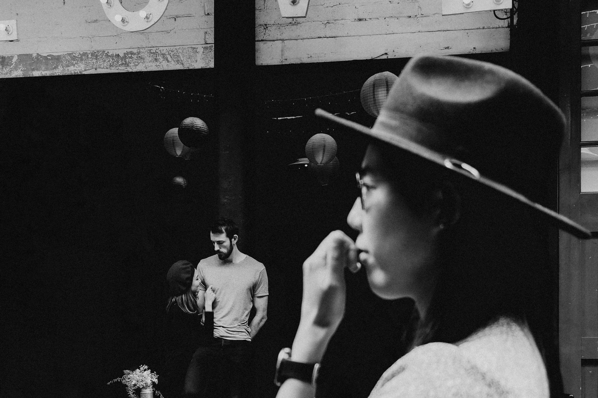 A black and white photo of a woman wearing a hat and glasses in the foreground, with a man and woman in the background, the woman wearing a beanie and the man smiling and looking down at a wedding event in london. Shot by Jason Evans photography Brid