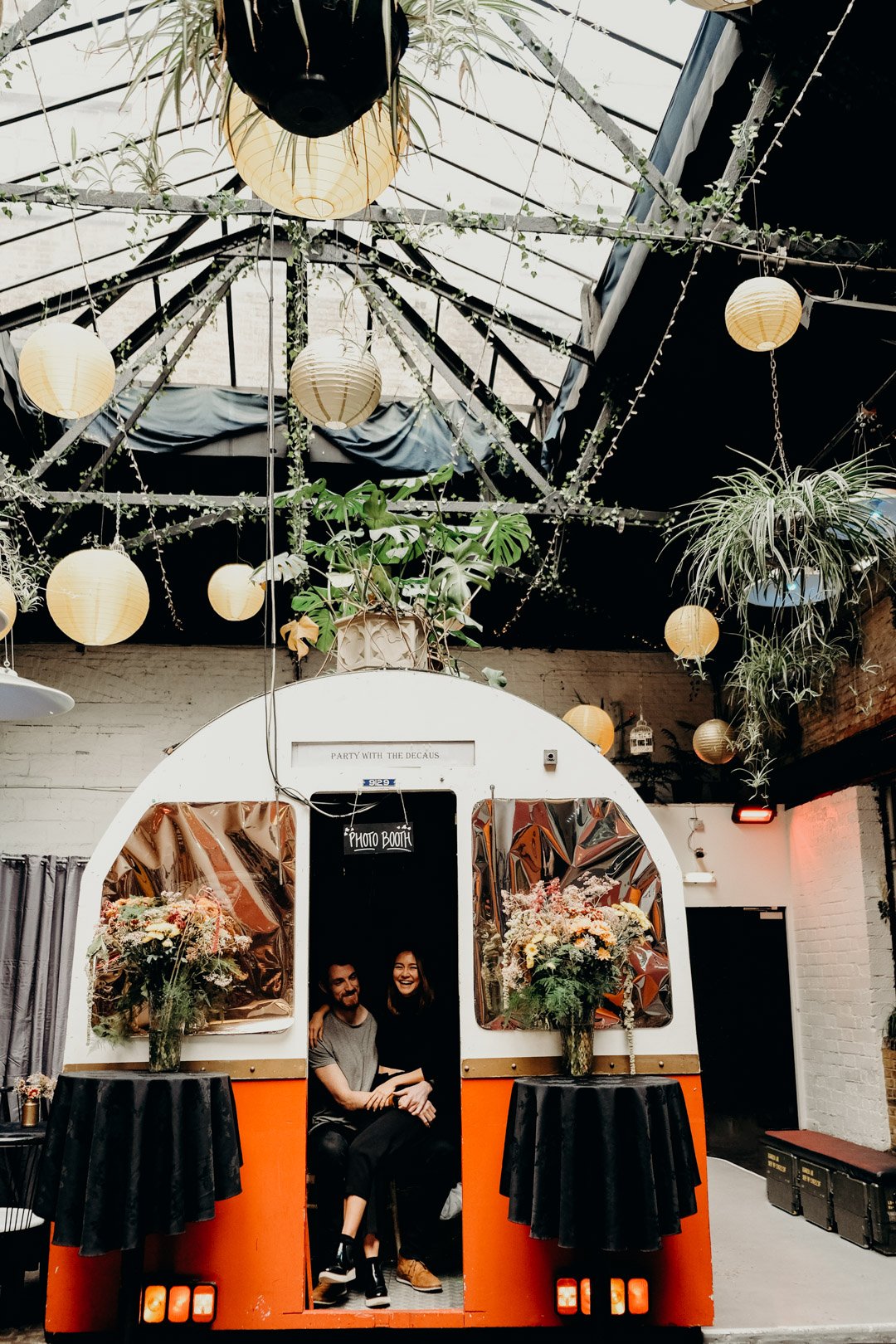 A photo booth inside a decorated indoor space with hanging paper lanterns, plants, and flowers, featuring two people sitting inside and smiling. Shot by Jason Evans photography Bridgend