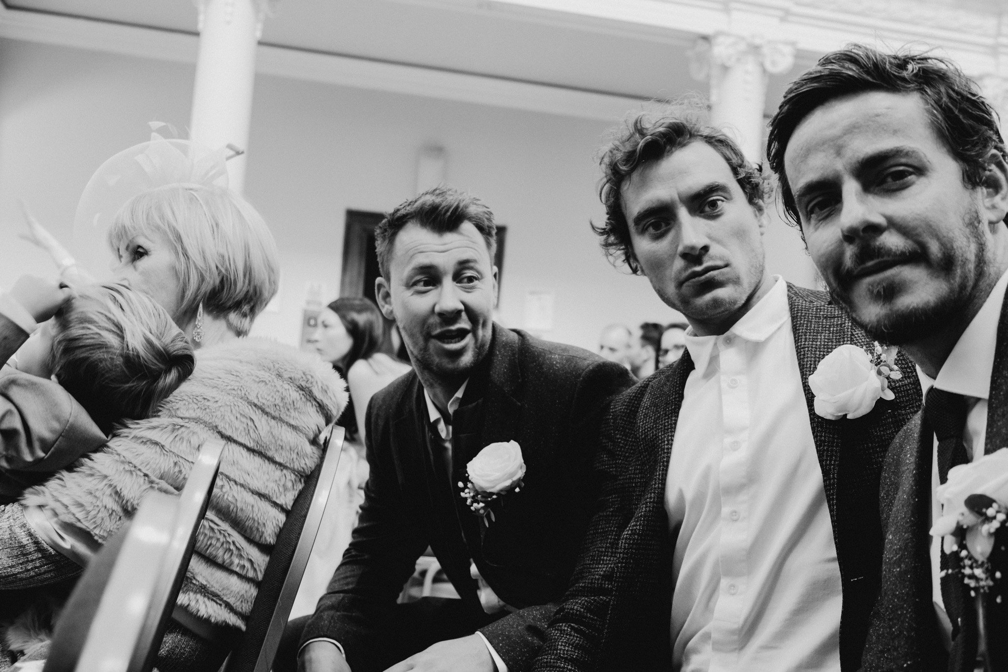 Three men seated at a wedding dressed in suits with boutonnieres, looking at the camera, with women in the background, indoors. Shot by Jason Evans photography Bridgend