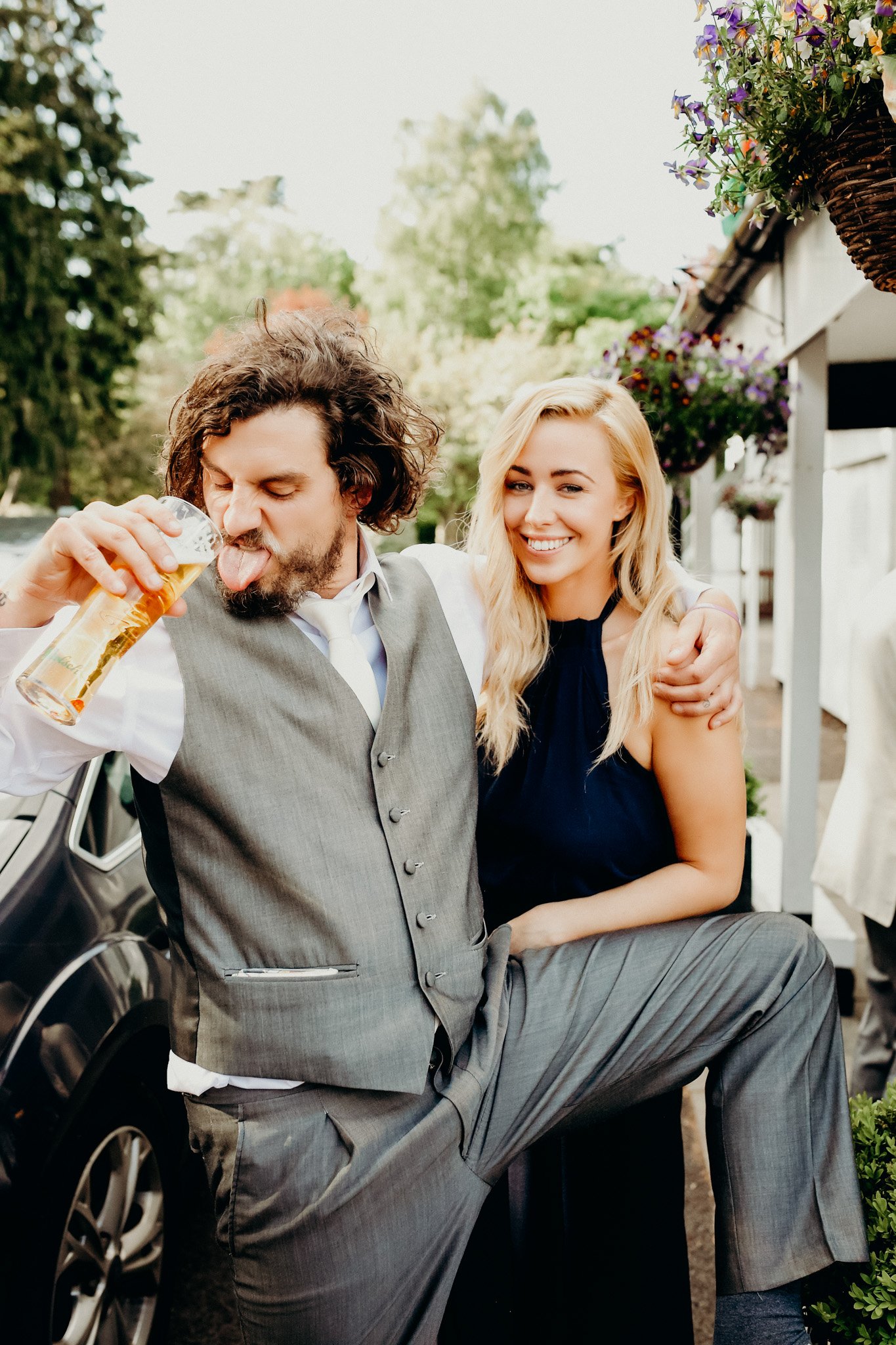 A man with curly hair and a beard, wearing a gray vest and pants, is drinking beer while posing with a woman with long blonde hair, wearing a dark blue dress, at a wedding in bridgend. The woman is smiling and has her arm around the man's shoulders a