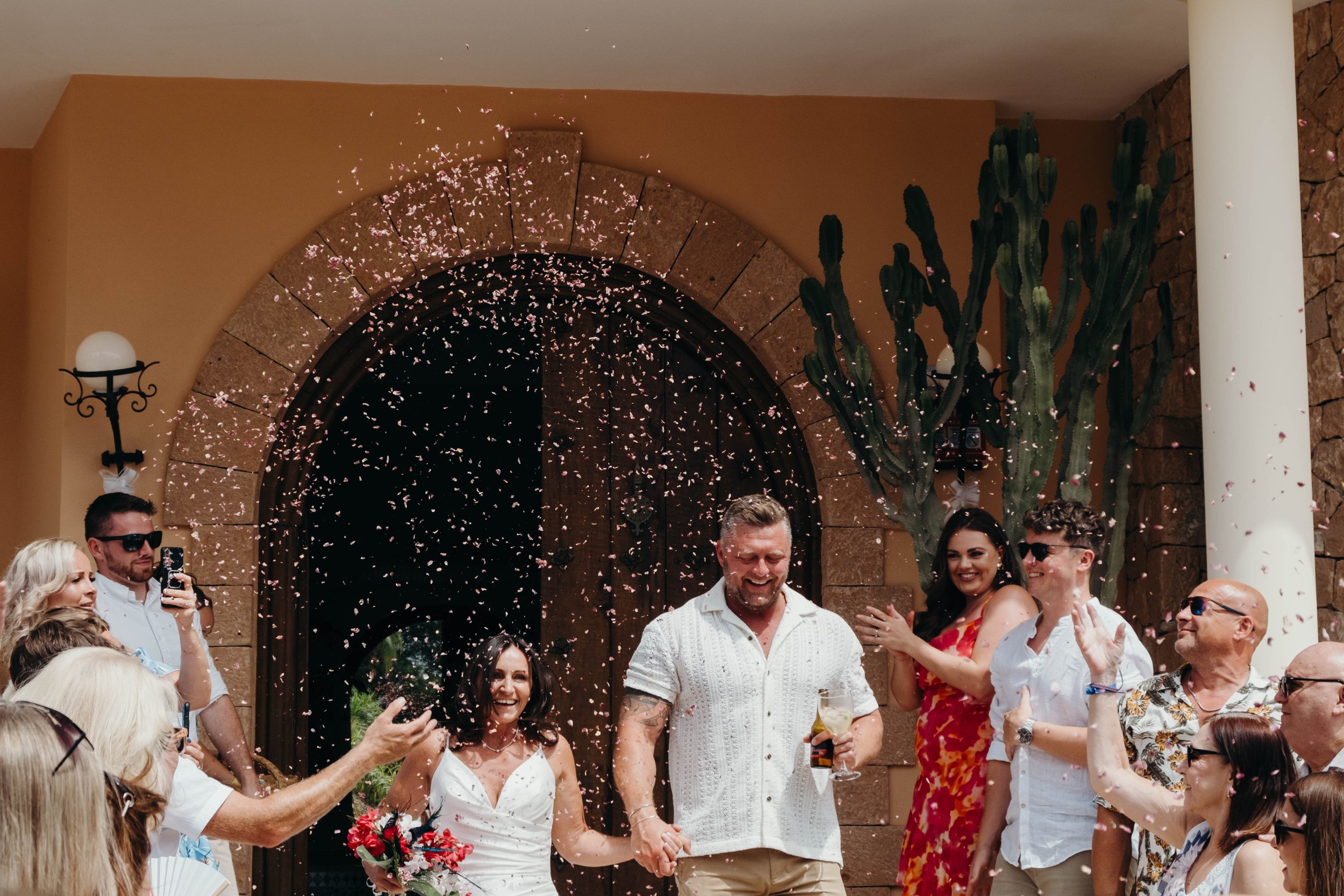 A group of people celebrating a wedding in Ibiza, outside a building with a large arched door, some wearing sunglasses, and pink flower petals falling around, with cacti and stone walls in the background. Shot by Jason Evans photography Bridgend