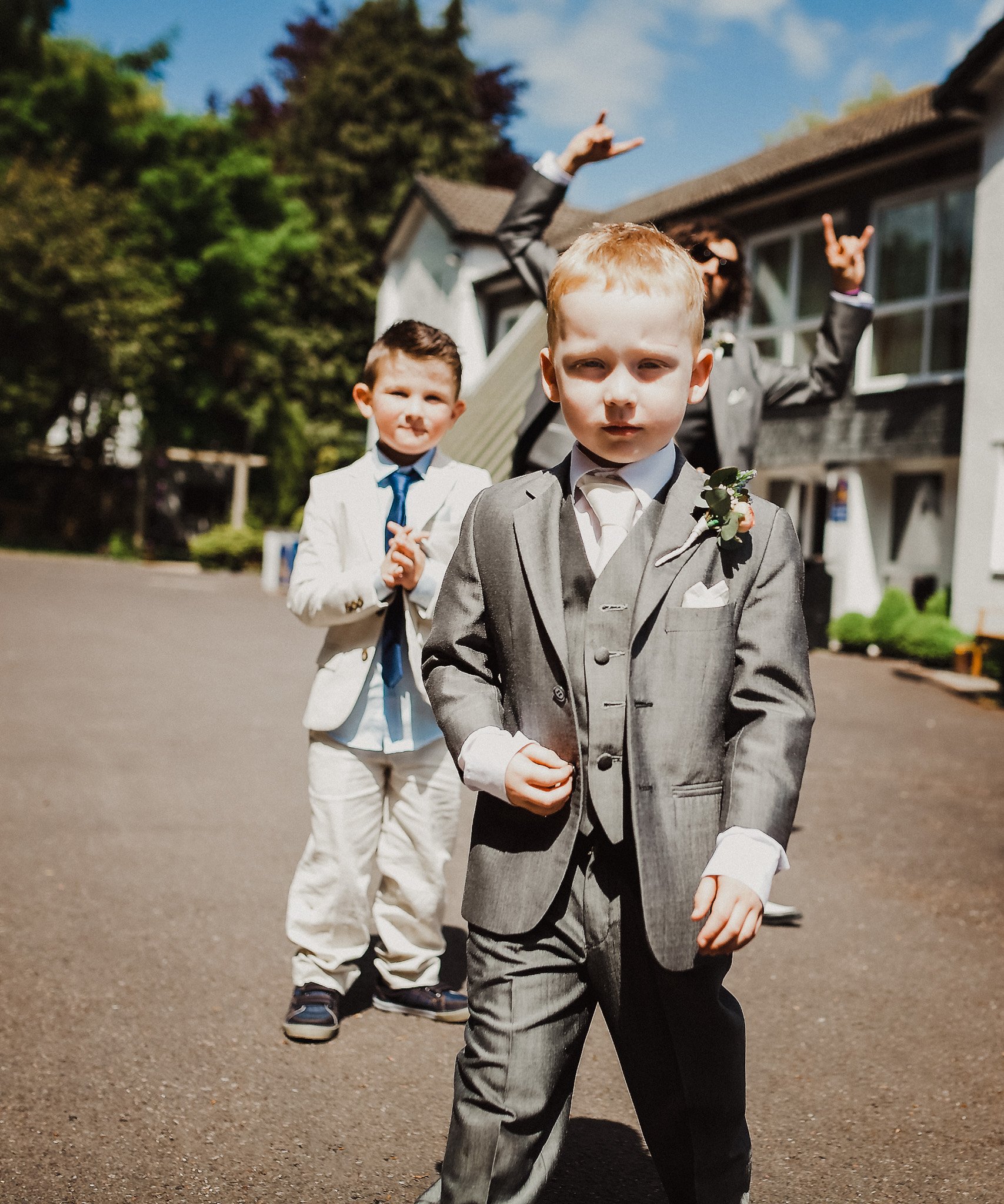 Three young boys dressed in suits outside on a sunny day, with a woman behind them making a rock sign with her hands. The boys appear to be at a wedding in Bridgend. Shot by Jason Evans photography Bridgend