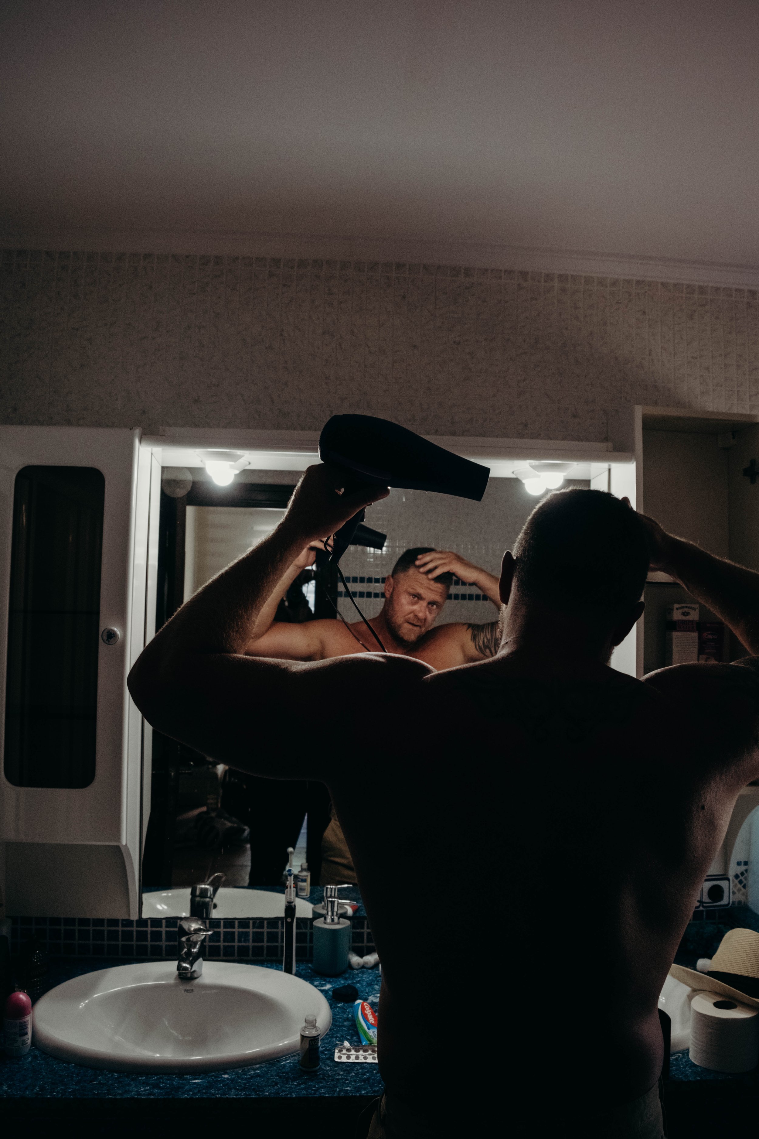 A shirtless man with tattoos looks into a mirror while drying his hair with a hairdryer in a bathroom, illuminated by vanity lights above the mirror at a wedding in Ibiza, spain. Shot by Jason Evans photography Bridgend