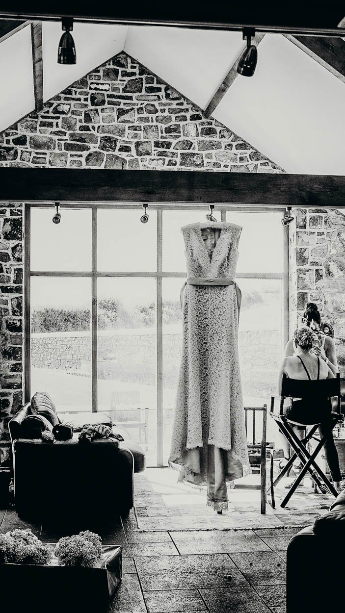A wedding dress hanging in front of large windows with a rustic brick wall interior. Several people are sitting on chairs, and there are lounge furniture and flowers inside. Shot by Jason Evans photography Bridgend