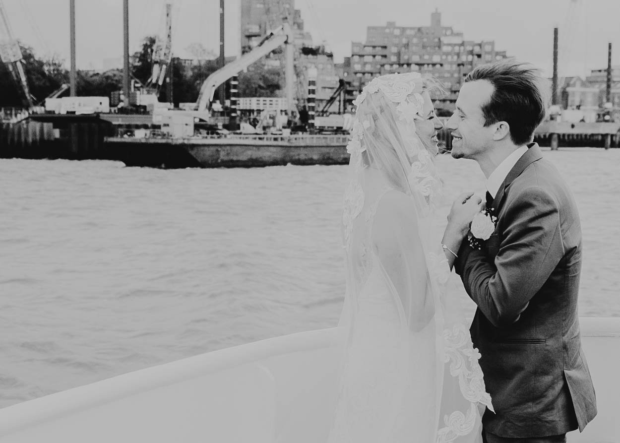 A bride and groom share a moment by the water with a harbor and bridge in the background in London. The black-and-white photo captures their happy expressions. Shot by Jason Evans photography Bridgend