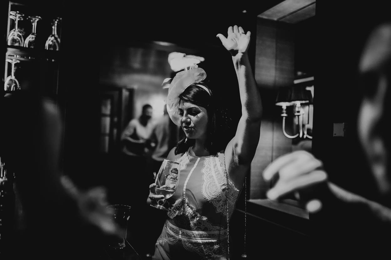 A woman in a lace dress with a head wrap dancing with her hand raised in a bar or nightclub setting, surrounded by blurred figures and barware at a wedding in bridgend, South Wales. Shot by Jason Evans photography Bridgend